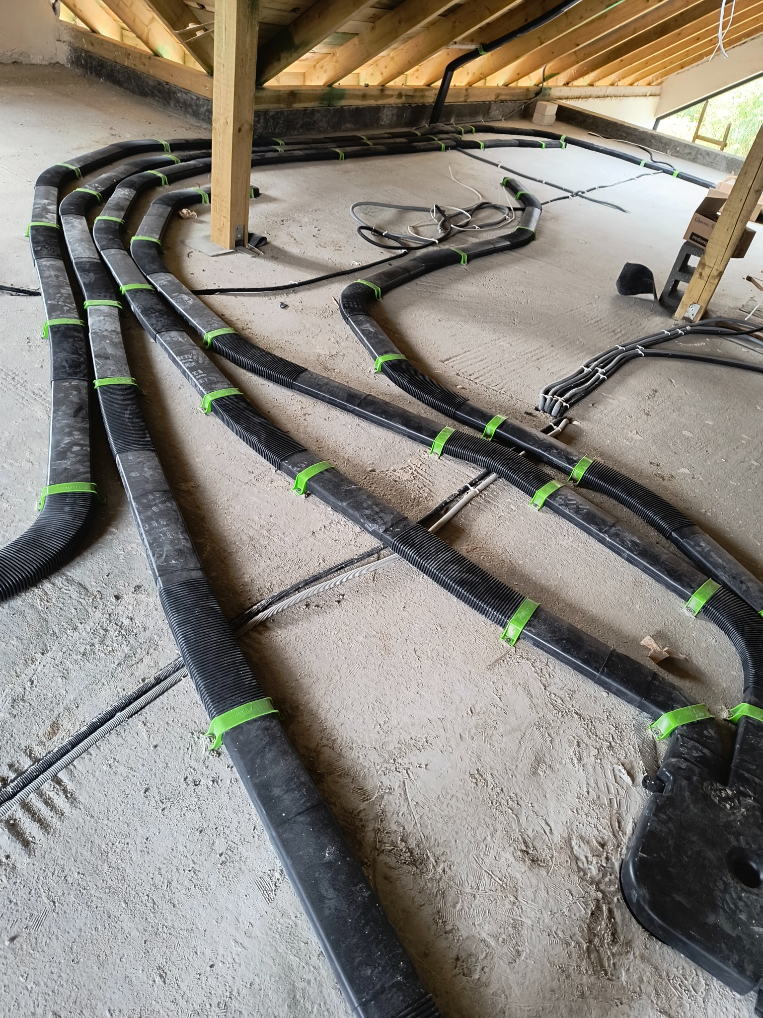 Czarny system rur wentylacyjnych, zabezpieczony zielonymi klamrami, rozłożony na surowym podłożu w trakcie instalacji pod drewnianą konstrukcją dachu.