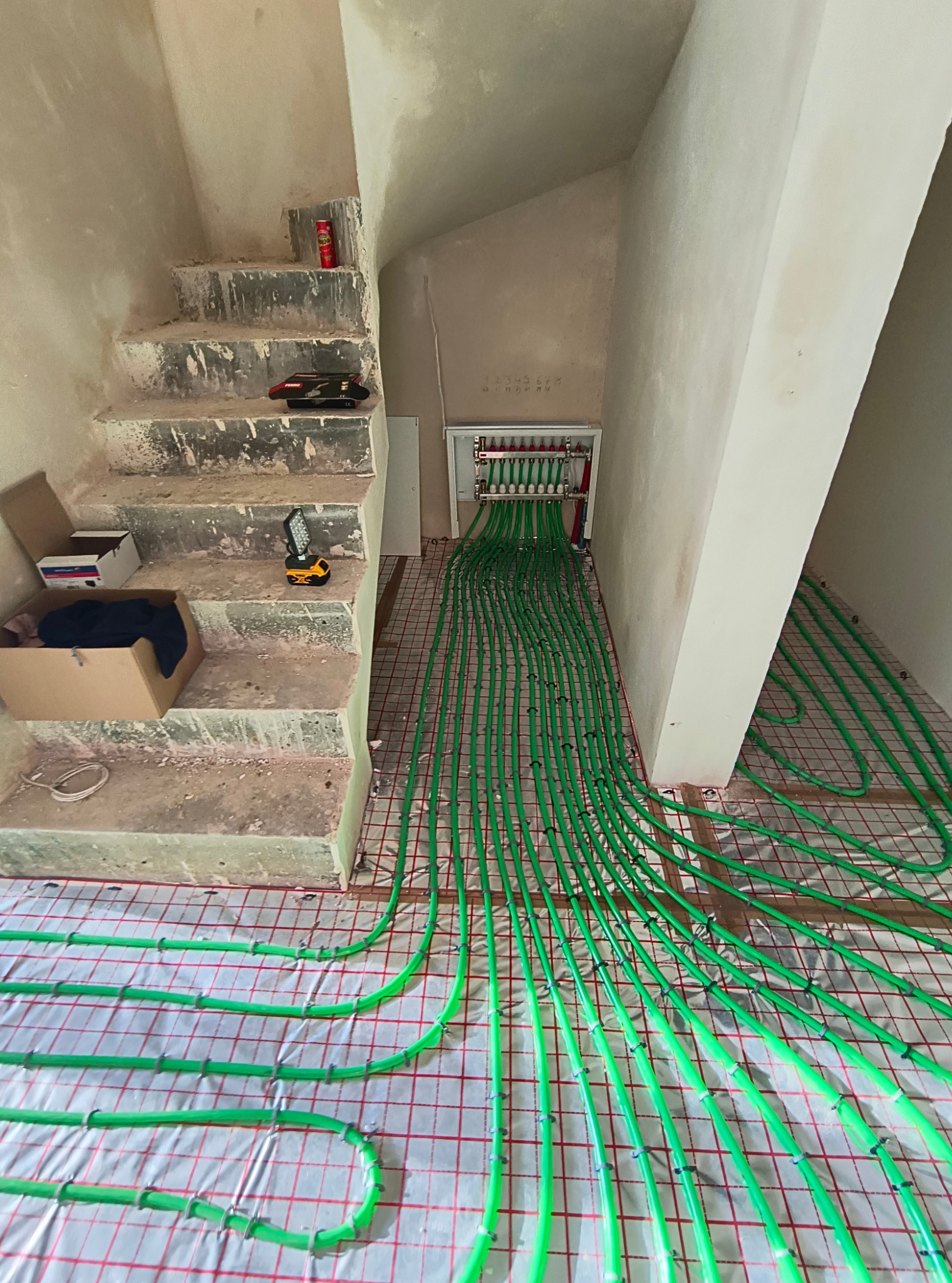 Zielone rury ogrzewania podłogowego ułożone na folii z siatką, prowadzące do rozdzielacza zamontowanego na ścianie w surowym wnętrzu klatki schodowej, widoczne narzędzia i materiały budowlane...