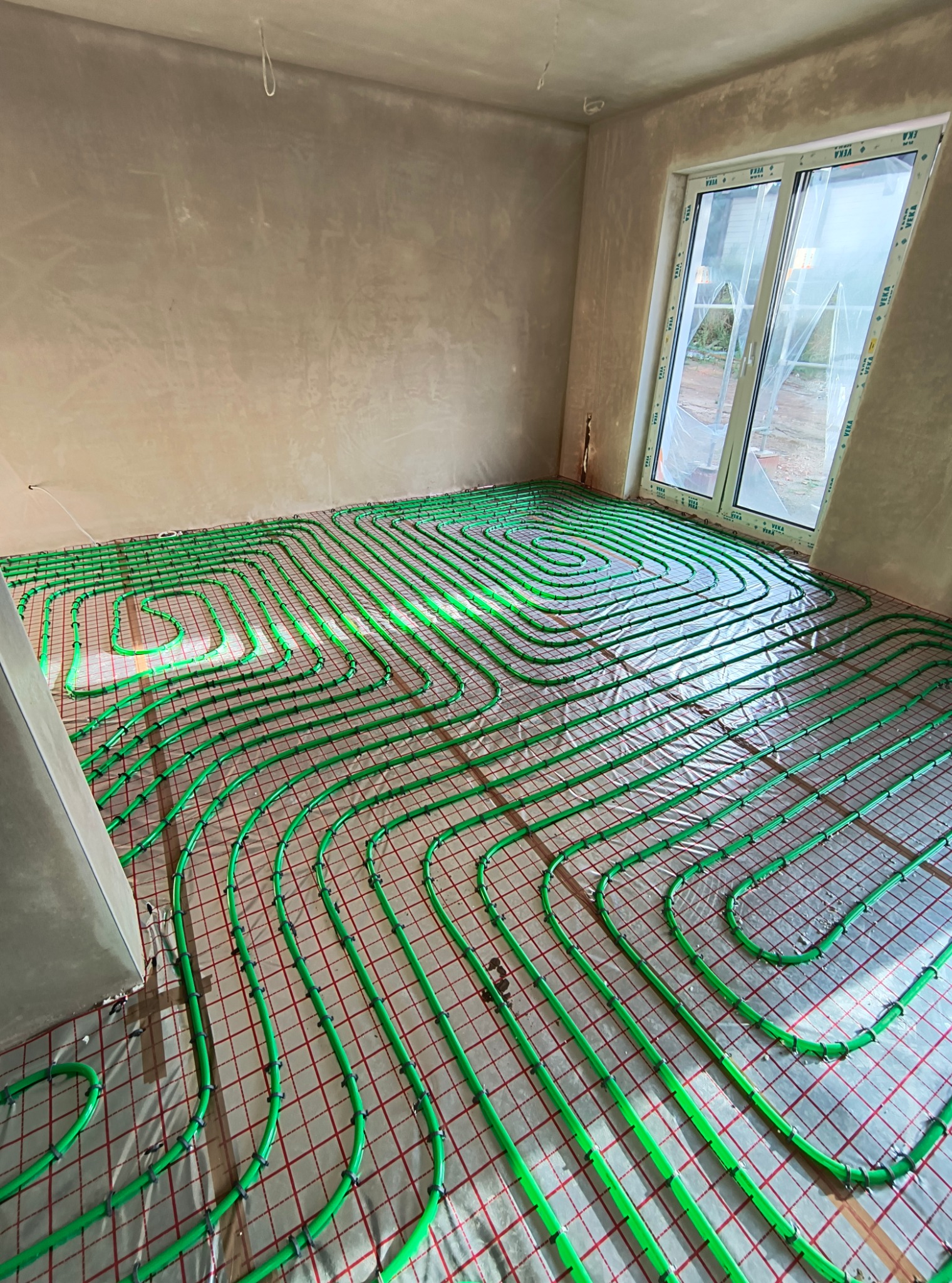 Zielone rury ogrzewania podłogowego ułożone na czerwonej siatce w surowym pomieszczeniu z dużym oknem, widoczne niedokończone ściany.