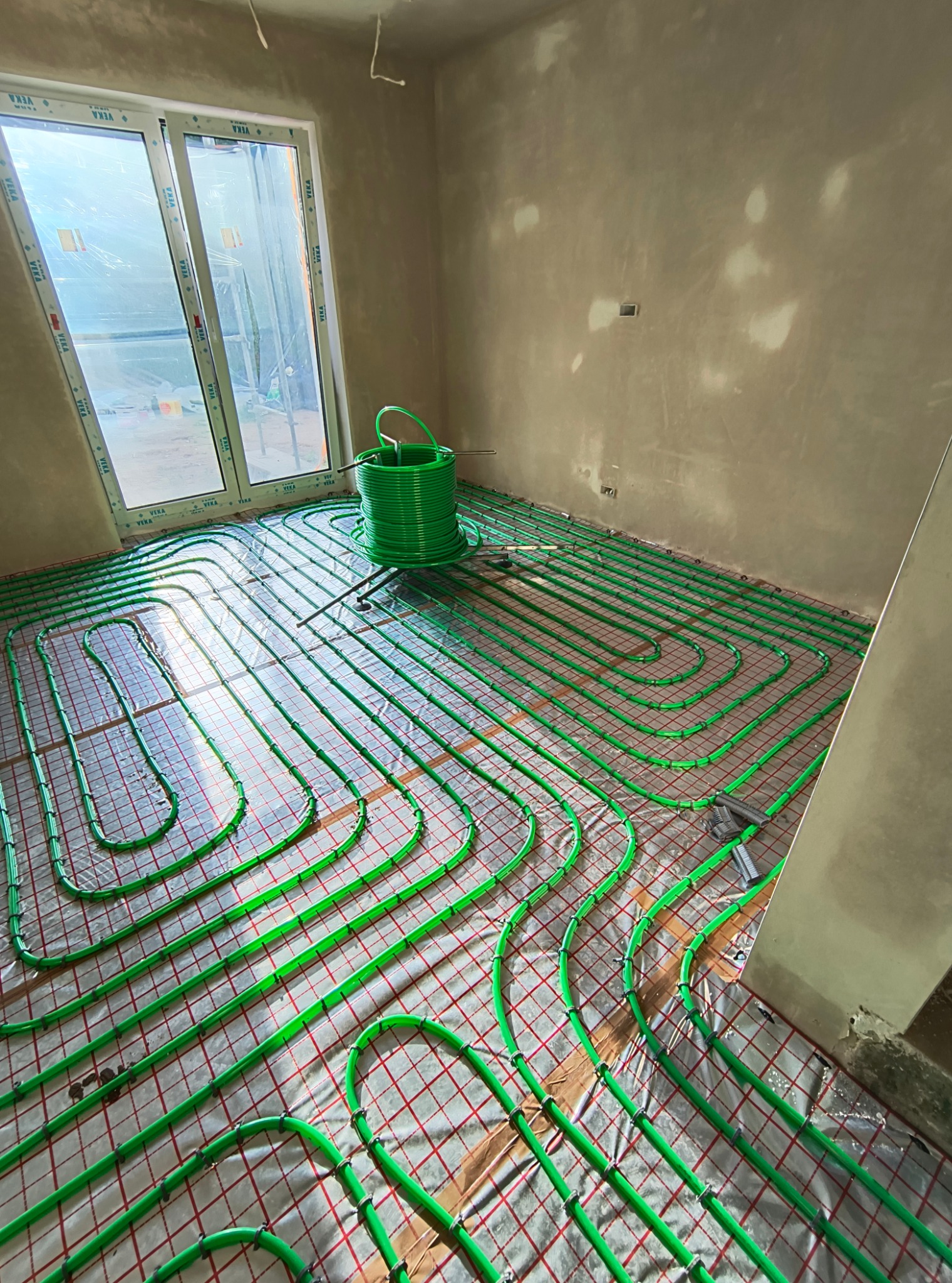 Instalacja ogrzewania podłogowego z zielonych rurek na folii z czerwoną siatką, widoczne okno z folią ochronną i ściany w stanie surowym.