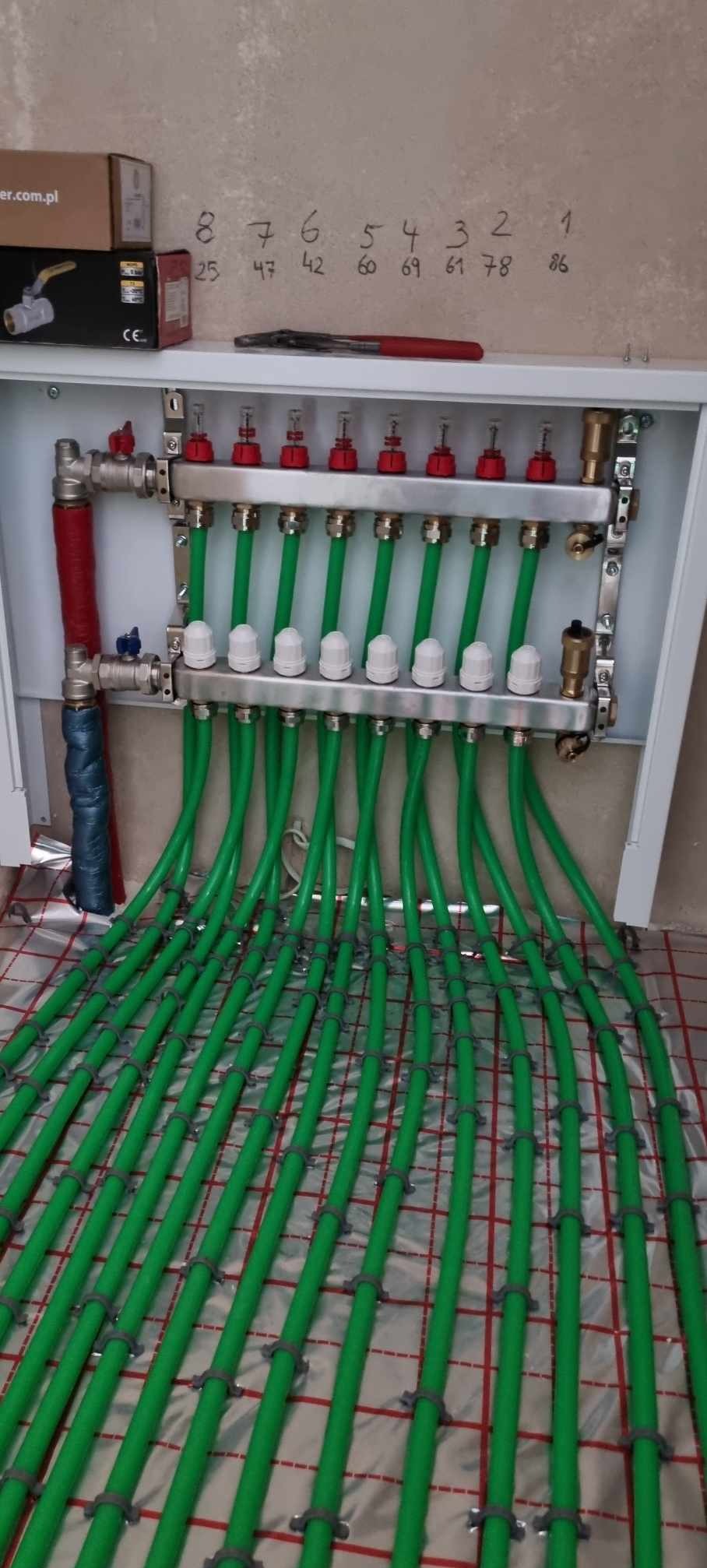 Rozdzielacz ogrzewania podłogowego z zielonymi rurami na folii aluminiowej z siatką, widoczne zawory i numeracja rur na ścianie.