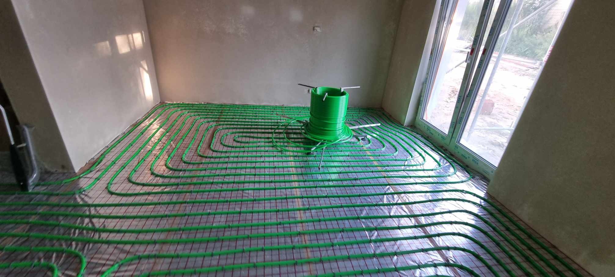 Zielone rury ogrzewania podłogowego ułożone na folii odblaskowej z widoczną rolką rur w surowym pomieszczeniu z oknem.