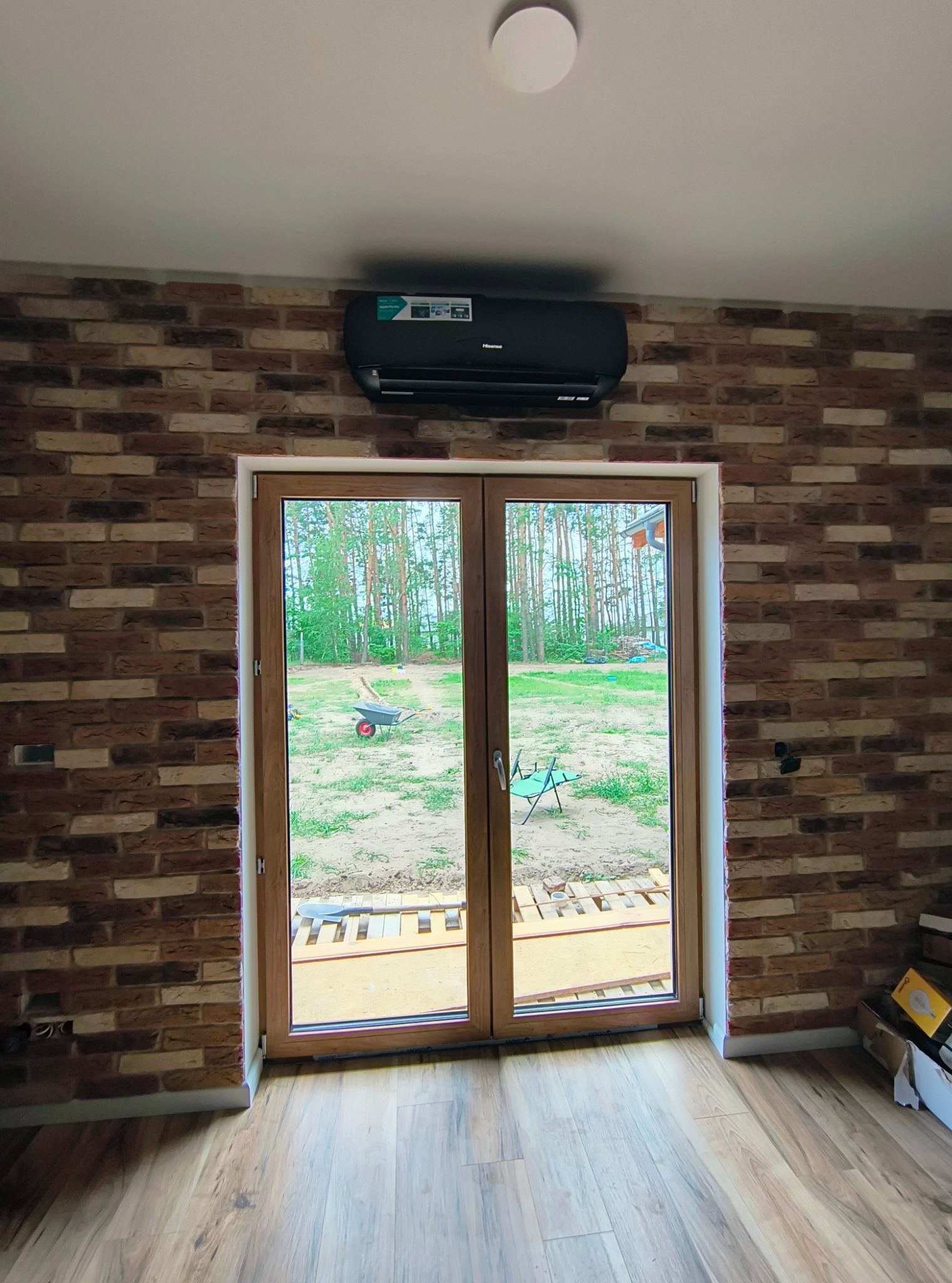 Czarny klimatyzator ścienny zamontowany nad drewnianymi drzwiami balkonowymi w pokoju z ceglaną ścianą, widok na zewnątrz na trawnik z taczką.