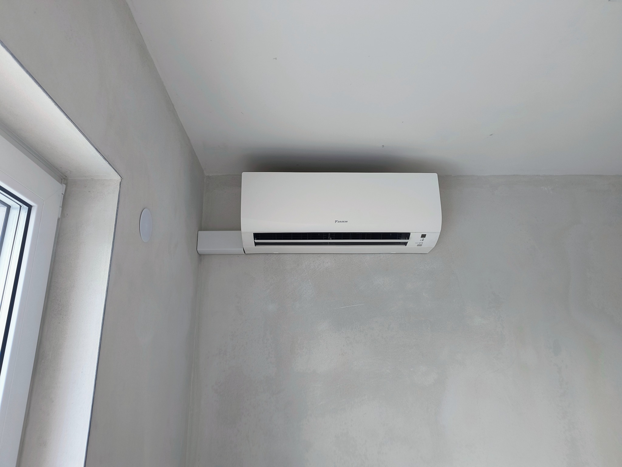 Biała jednostka klimatyzacyjna Daikin zamontowana na surowej, szarej ścianie w nowym budynku, widoczny fragment okna po lewej stronie.