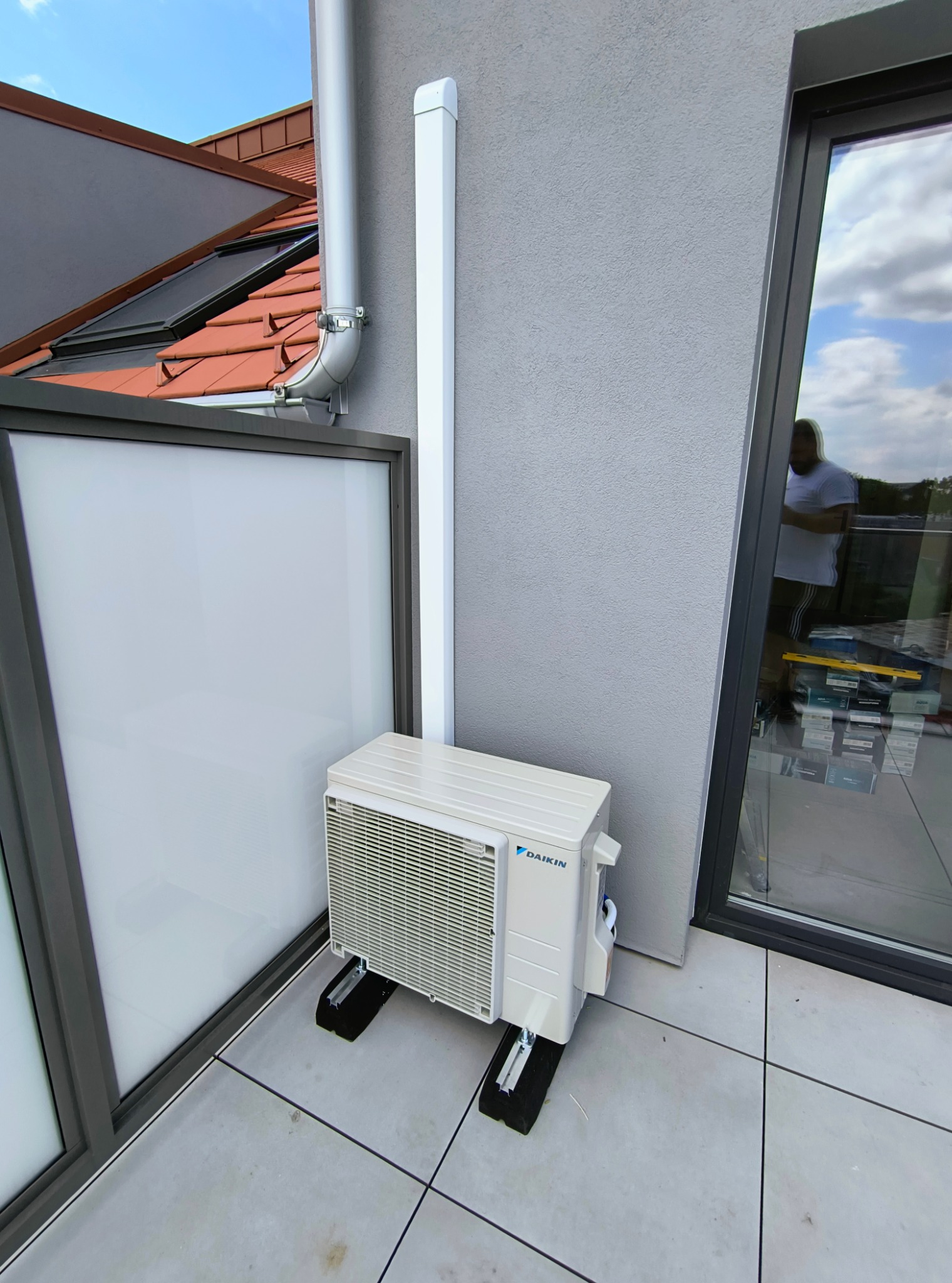 Biała jednostka zewnętrzna klimatyzacji Daikin zamontowana na balkonie na specjalnych wspornikach, z widoczną rurą odprowadzającą skropliny, na tle szarej ściany budynku i okna z odbiciem