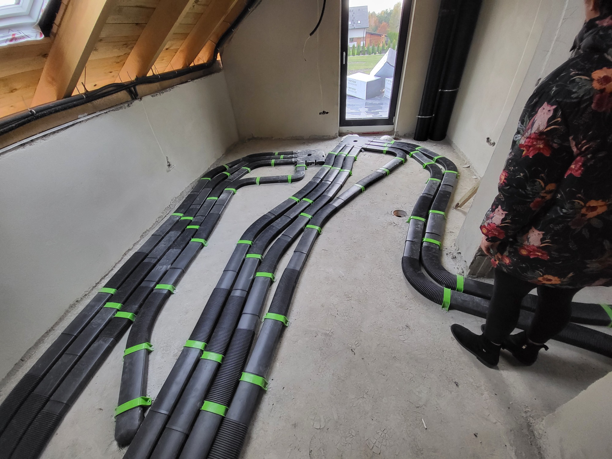 Rozprowadzenie czarnych, elastycznych rur wentylacyjnych z zielonymi obejmami na poddaszu w trakcie budowy, widok z góry, fragment sylwetki osoby w kwiecistej bluzce.