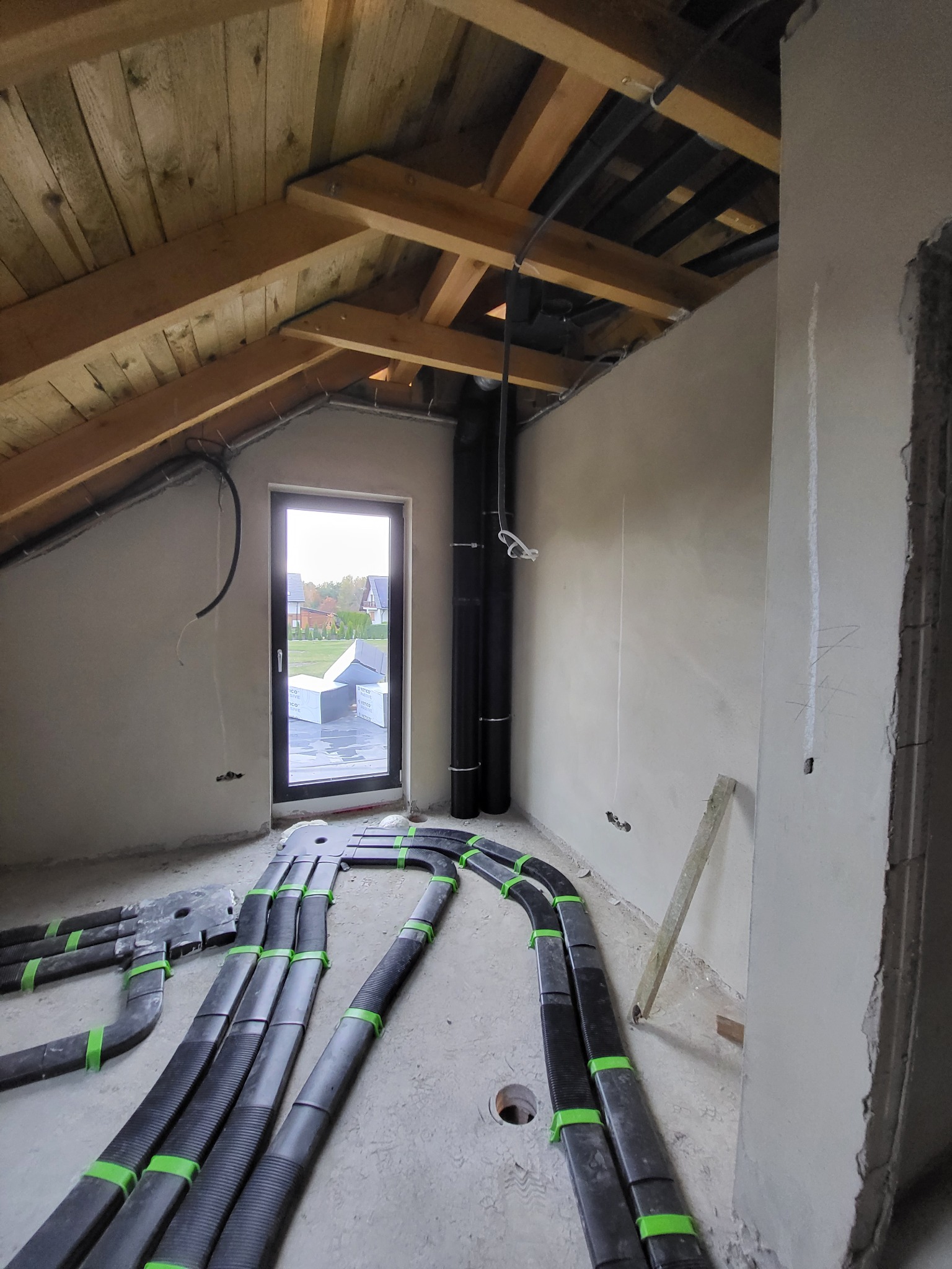 Surowy stan poddasza z widocznymi czarnymi rurami wentylacyjnymi z zielonymi opaskami ułożonymi na podłodze, pionowe rury przy ścianie, drewniana konstrukcja dachu i okno z widokiem na ogród.