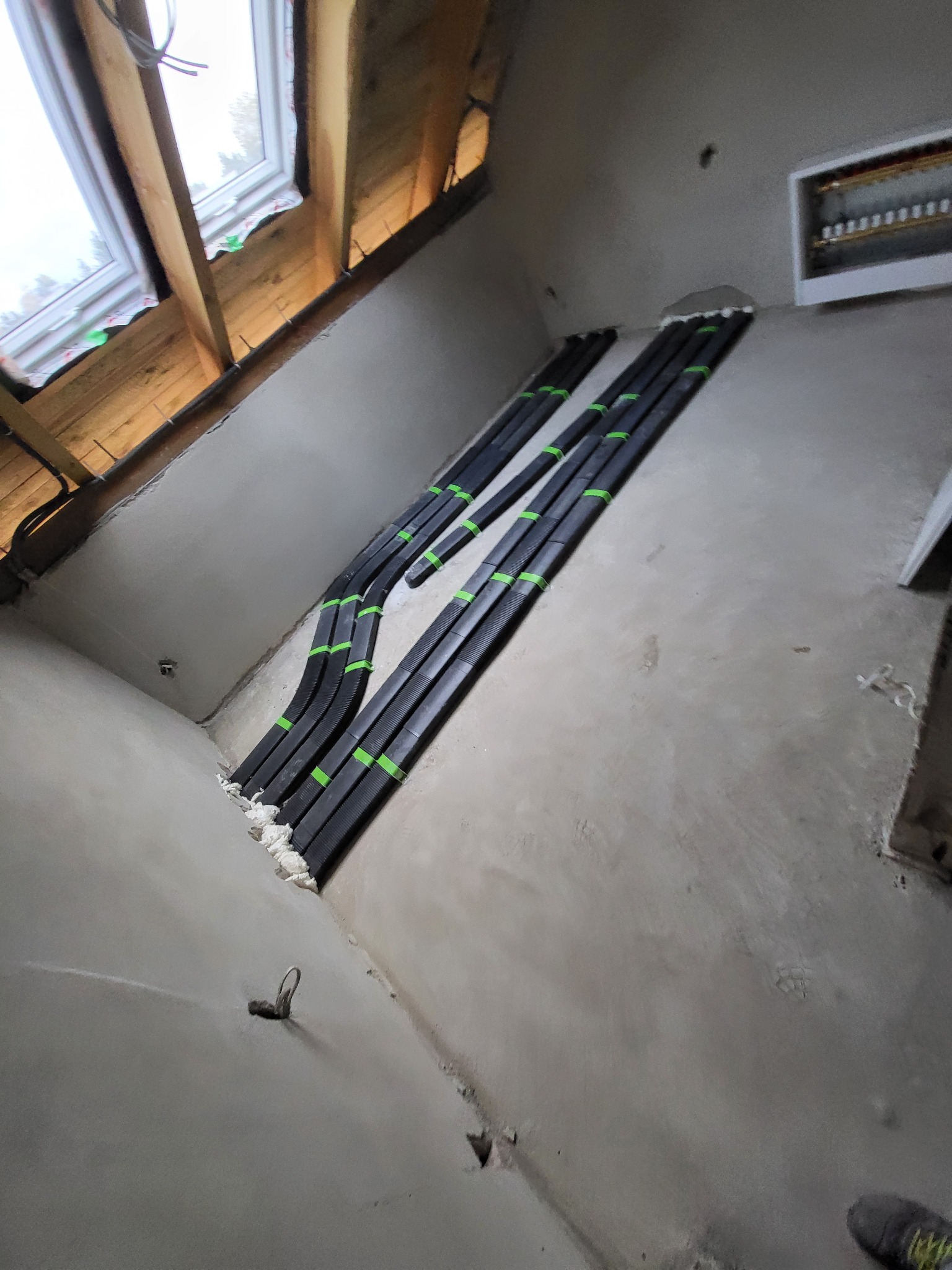 Cztery czarne rury wentylacyjne zabezpieczone zieloną taśmą, prowadzone po szarej ścianie pod skosem, z widocznym oknem dachowym w tle.