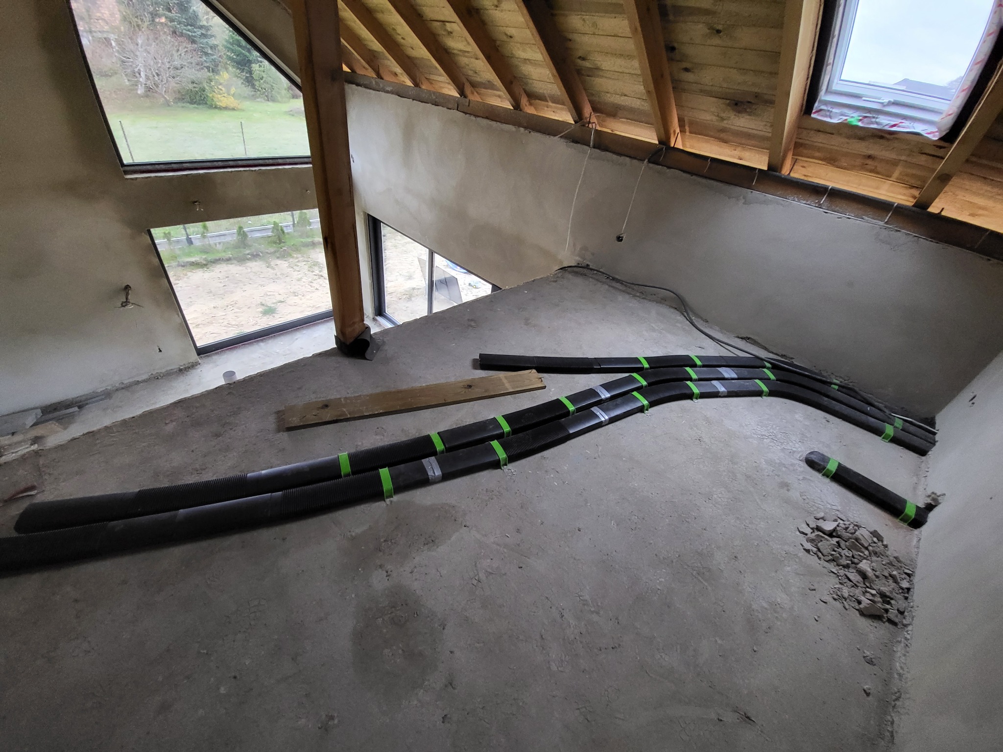 Wnętrze w stanie surowym z widoczną instalacją wentylacyjną z czarnych rur spiętych zieloną taśmą, drewniana konstrukcja dachu, okno dachowe i duże okno z widokiem na ogród.