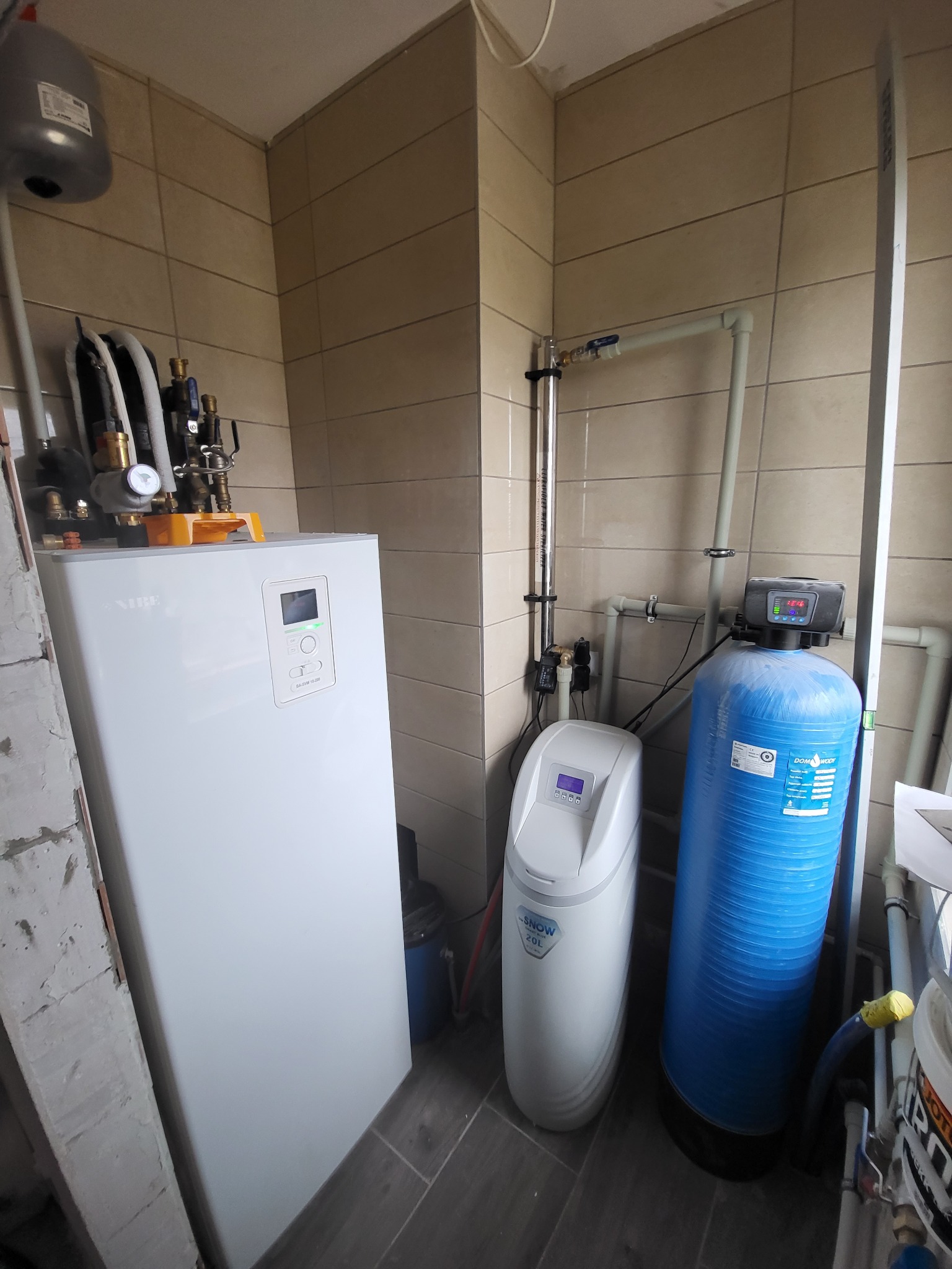 Biały piec grzewczy Nibe, obok zmiękczacz wody Snow 20L oraz niebieski filtr wody Domo Body, na tle beżowych płytek, widoczne rury instalacji.