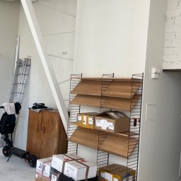 ADizajn - Białe wnętrze w trakcie prac, z widocznymi rurami wentylacyjnymi pod sufitem, drewnianymi półkami String z kartonami, drabiną opartą o ścianę i hulajnogą elektryczną w tle, oraz stertą zapakowanych...