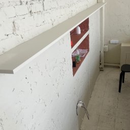 ADizajn - Biała ściana z cegły i tynku z wnęką, w której znajdują się dekoracyjny dyfuzor zapachowy, butelka z napojem i dokumenty, widoczny fragment podłogi i krzesła.