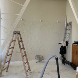 ADizajn - Wnętrze w trakcie prac wykończeniowych: dwie drabiny, wentylator, odkurzacz przemysłowy, wiadro farby stoją na podłodze, ściany z dekoracyjną fakturą, widoczne instalacje pod sufitem.