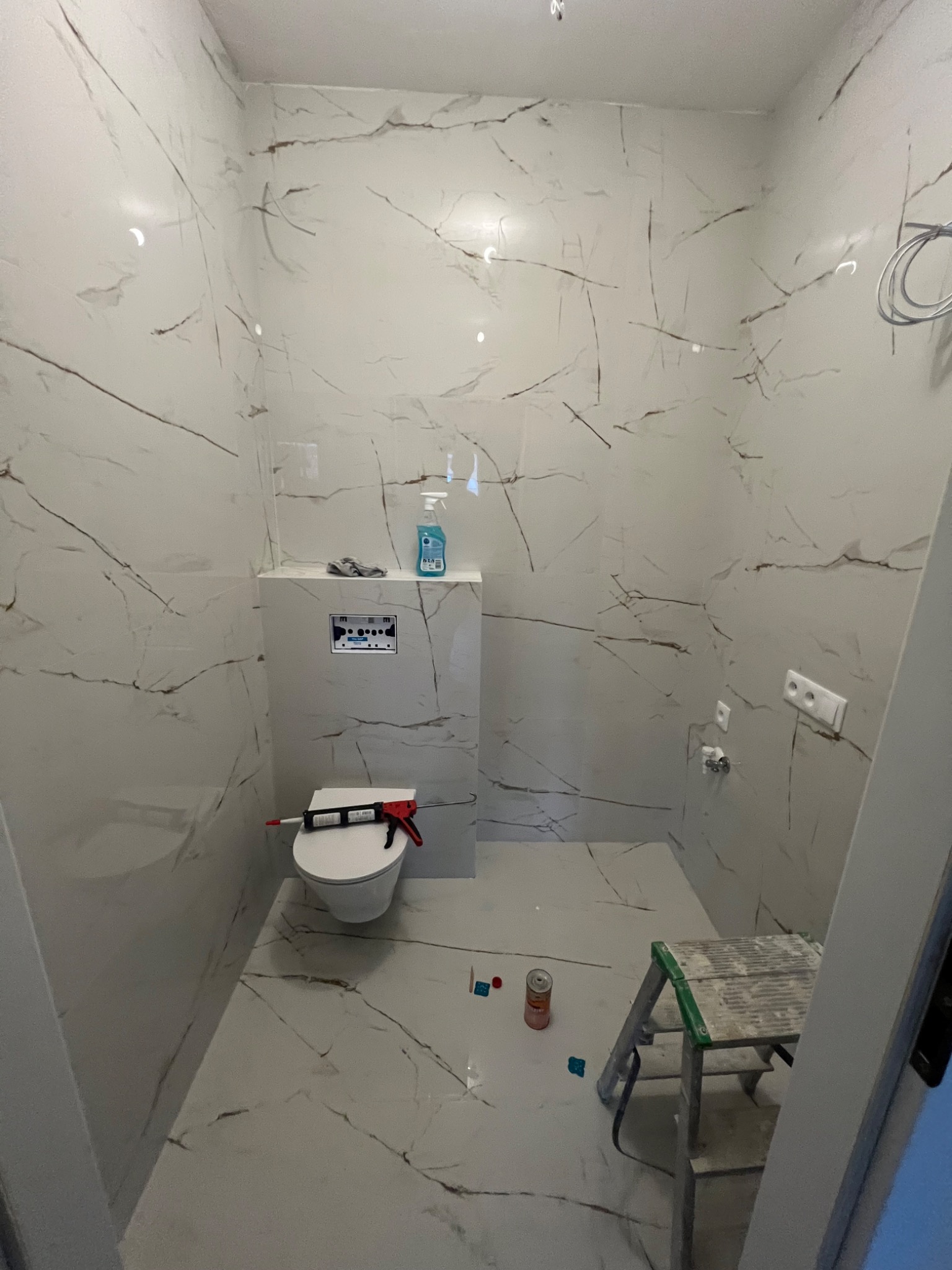 Wnętrze łazienki w trakcie wykańczania w Warszawie: białe płytki imitujące marmur na ścianach i podłodze, zamontowana toaleta z ukrytą spłuczką, pistolet do silikonu na sedesie, drabina malarska...