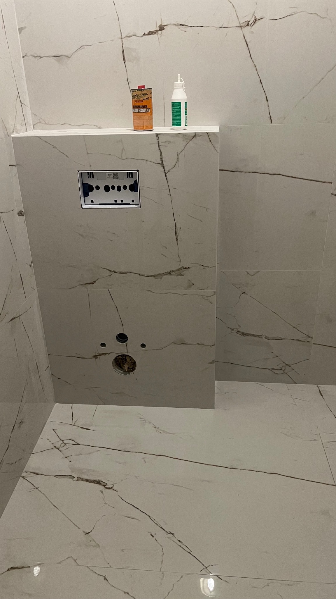 Białe marmurowe płytki w łazience, widoczne otwory na instalacje, puszka podtynkowa do WC, preparaty czyszczące na półce.