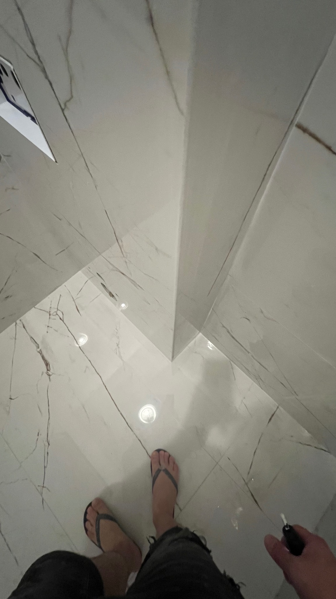 Białe, marmurowe płytki w łazience ułożone na podłodze i ścianach, widok z góry na stopy osoby w klapkach i rękę trzymającą czarny marker do fug