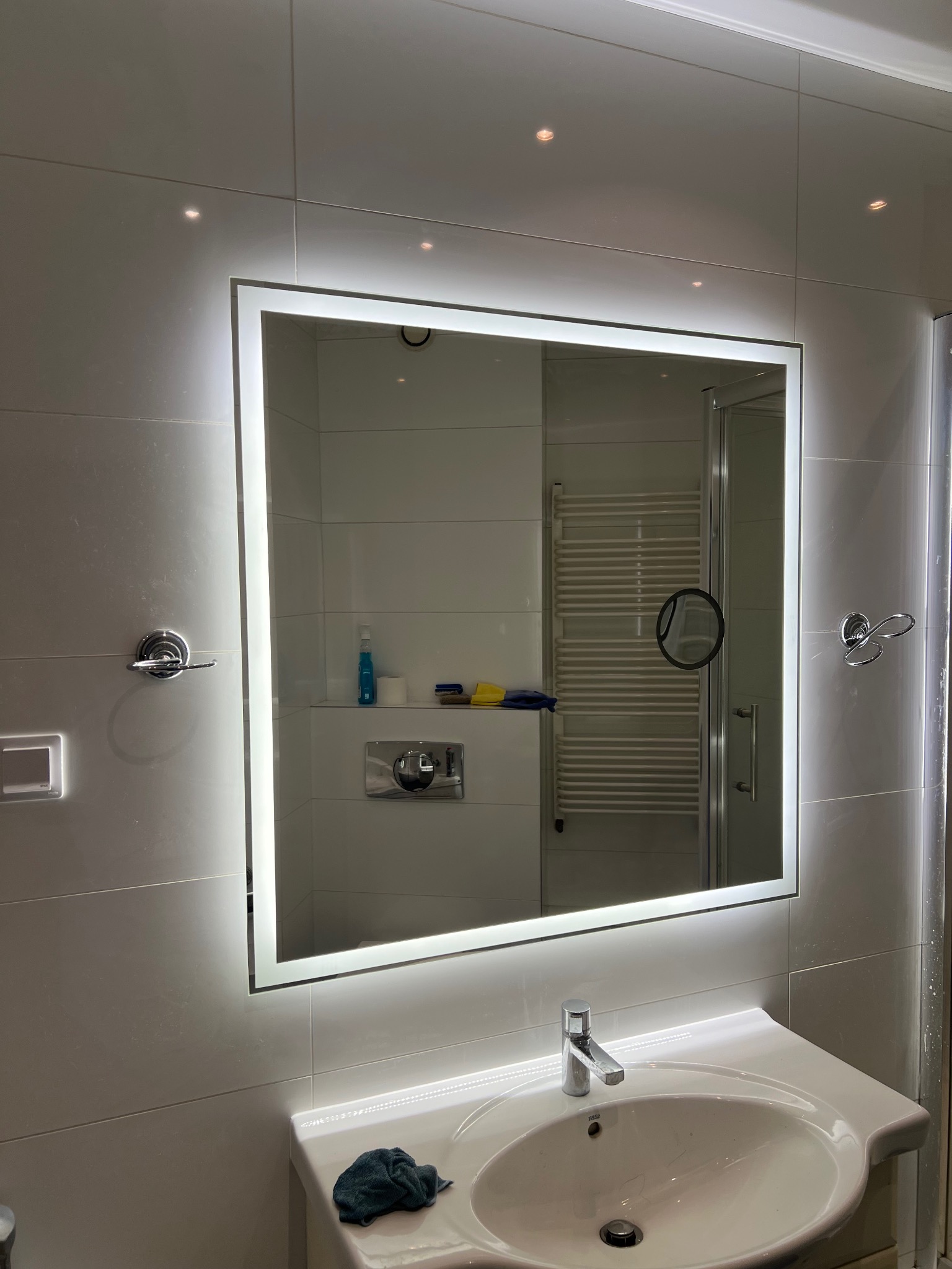 Nowoczesna łazienka z podświetlanym lustrem LED, białe płytki ścienne, umywalka z baterią, widoczny fragment kabiny prysznicowej i grzejnika drabinkowego.