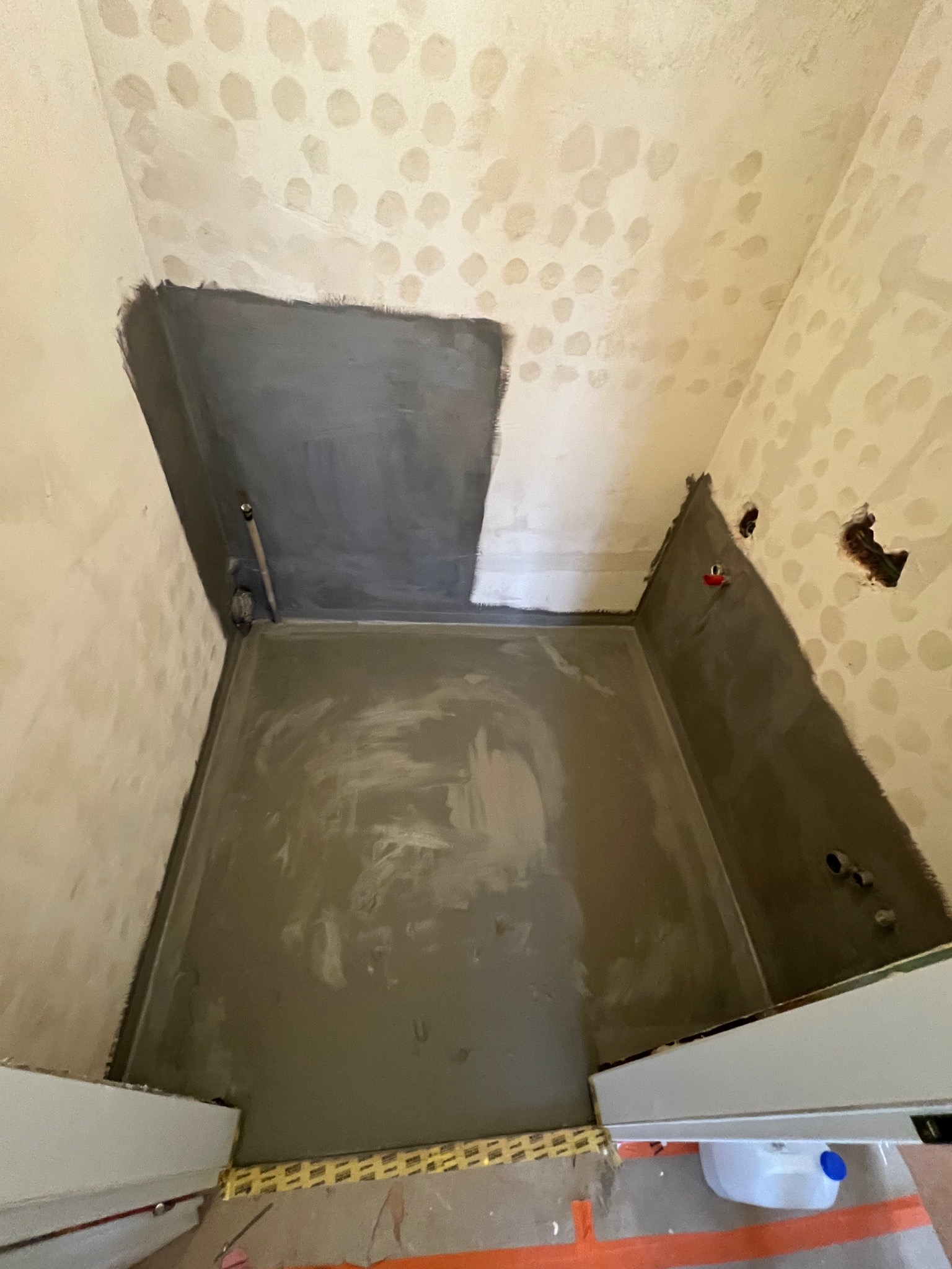 Przygotowanie do hydroizolacji w narożniku łazienki: gruntowanie ścian i wylewka na podłodze, widoczne wystające kable i rury.