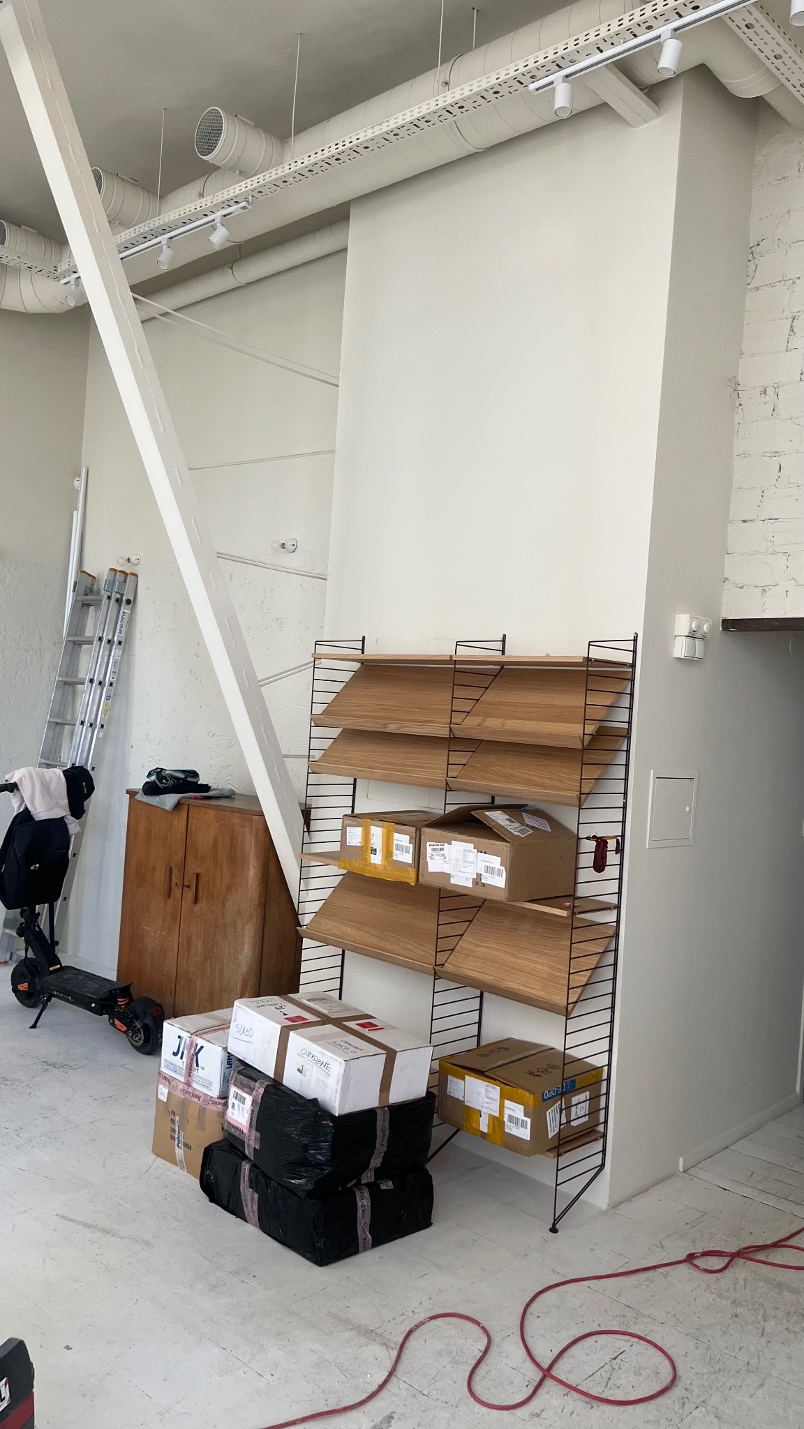 Białe wnętrze w trakcie prac, z widocznymi rurami wentylacyjnymi pod sufitem, drewnianymi półkami String z kartonami, drabiną opartą o ścianę i hulajnogą elektryczną w tle, oraz stertą zapakowanych...