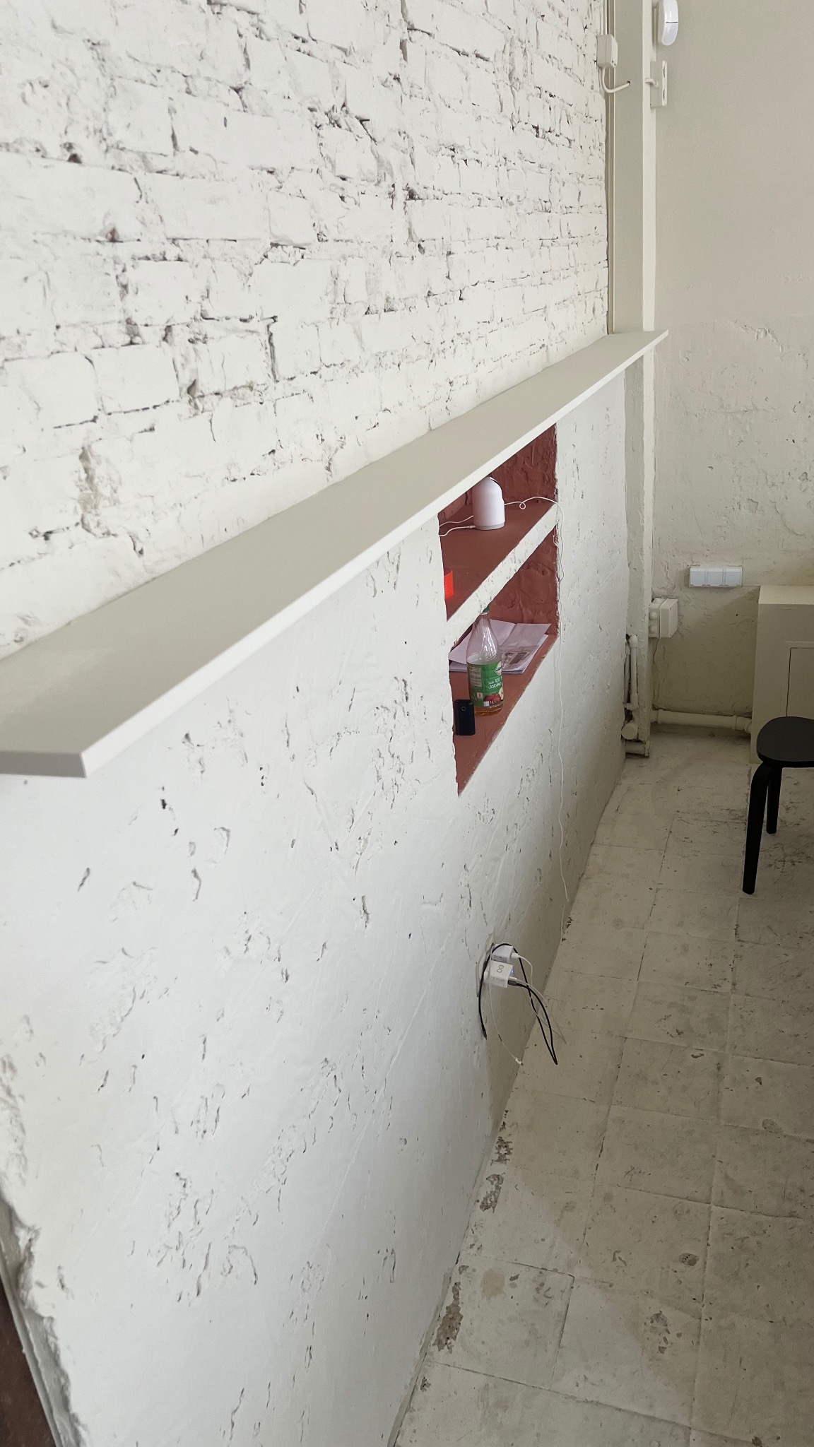 Biała ściana z cegły i tynku z wnęką, w której znajdują się dekoracyjny dyfuzor zapachowy, butelka z napojem i dokumenty, widoczny fragment podłogi i krzesła.