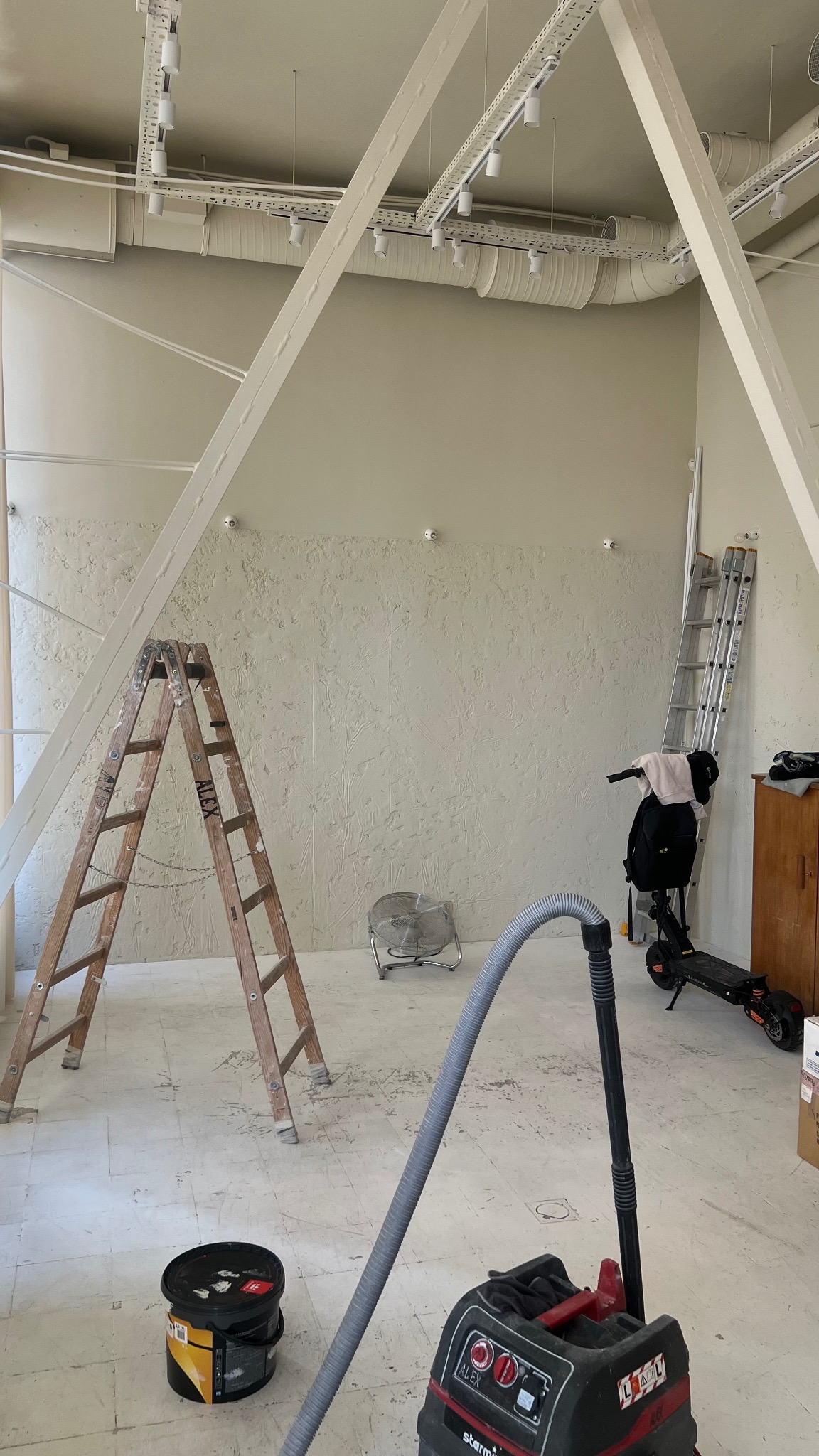Wnętrze w trakcie prac wykończeniowych: dwie drabiny, wentylator, odkurzacz przemysłowy, wiadro farby stoją na podłodze, ściany z dekoracyjną fakturą, widoczne instalacje pod sufitem.