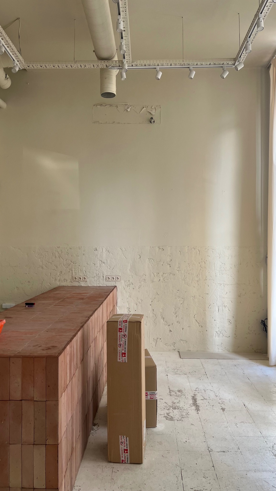 Wnętrze w trakcie prac wykończeniowych: murowany barek z cegieł, kartony z naklejkami ostrzegawczymi, ściana z widocznymi śladami po usuniętej tapecie oraz instalacja oświetleniowa na suficie.