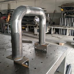 Pavlo Han - Metalowa konstrukcja w kształcie litery U, spawana do stalowych podstaw, leży na perforowanym stole warsztatowym; w tle iskry spawania i maszyny warsztatowe.