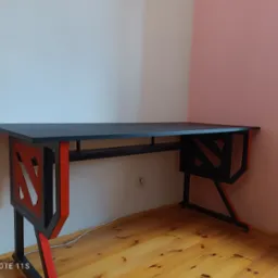 Czarno-czerwone biurko gamingowe z logo Dota 2 na nogach, ustawione w rogu pokoju z drewnianą podłogą i ścianami w odcieniach bieli i pudrowego różu.