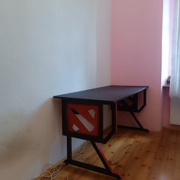 Pavlo Han - Czarno-czerwone biurko gamingowe z motywem graficznym na tle ściany w dwóch odcieniach różu i drewnianej podłogi.