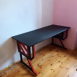 Pavlo Han - Czarno-czerwone biurko gamingowe z charakterystycznym wzorem na boku, stojące na drewnianej podłodze przy białej i różowej ścianie.