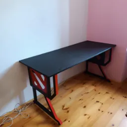 Czarno-czerwone biurko gamingowe z charakterystycznym wzorem na boku, stojące na drewnianej podłodze przy białej i różowej ścianie.