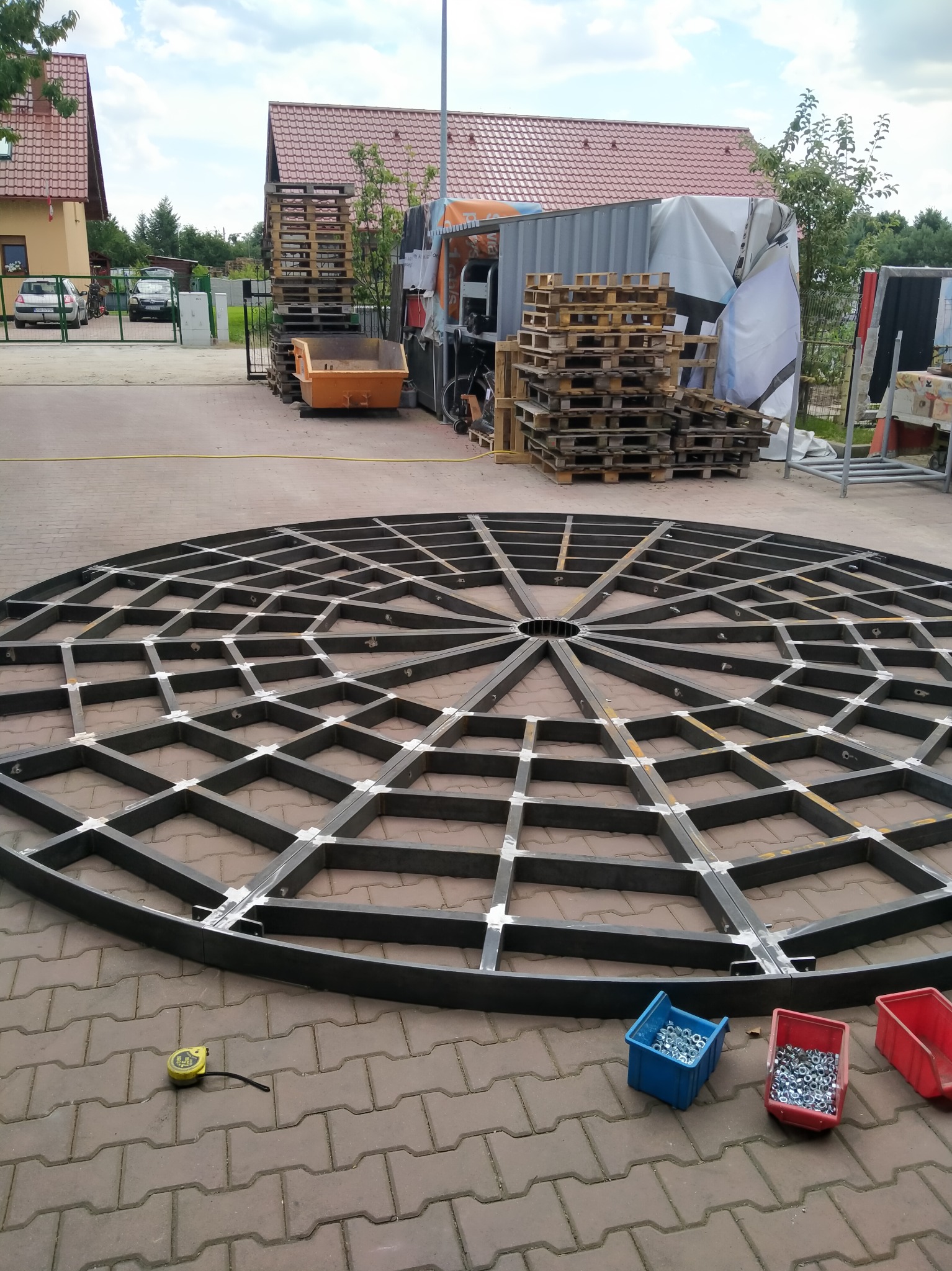 Metalowa konstrukcja okrągłej pergoli w trakcie budowy, widoczne spawy, śruby i miarka na kostce brukowej, w tle budynki i palety.