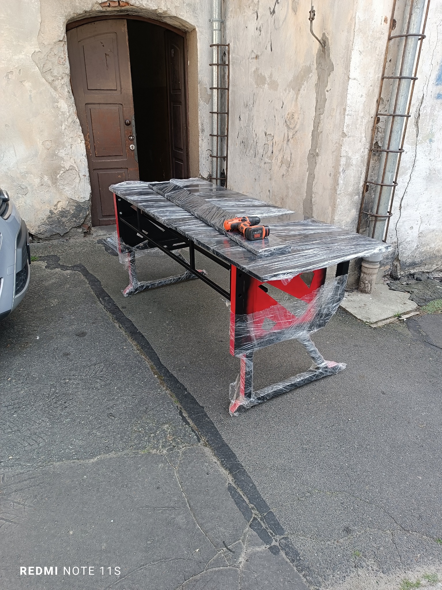 Czarny metalowy stół z czerwonymi akcentami, owinięty folią, stoi na zewnątrz przy starej, popękanej ścianie z rurami i otwartymi drzwiami. Na blacie leży wkrętarka.