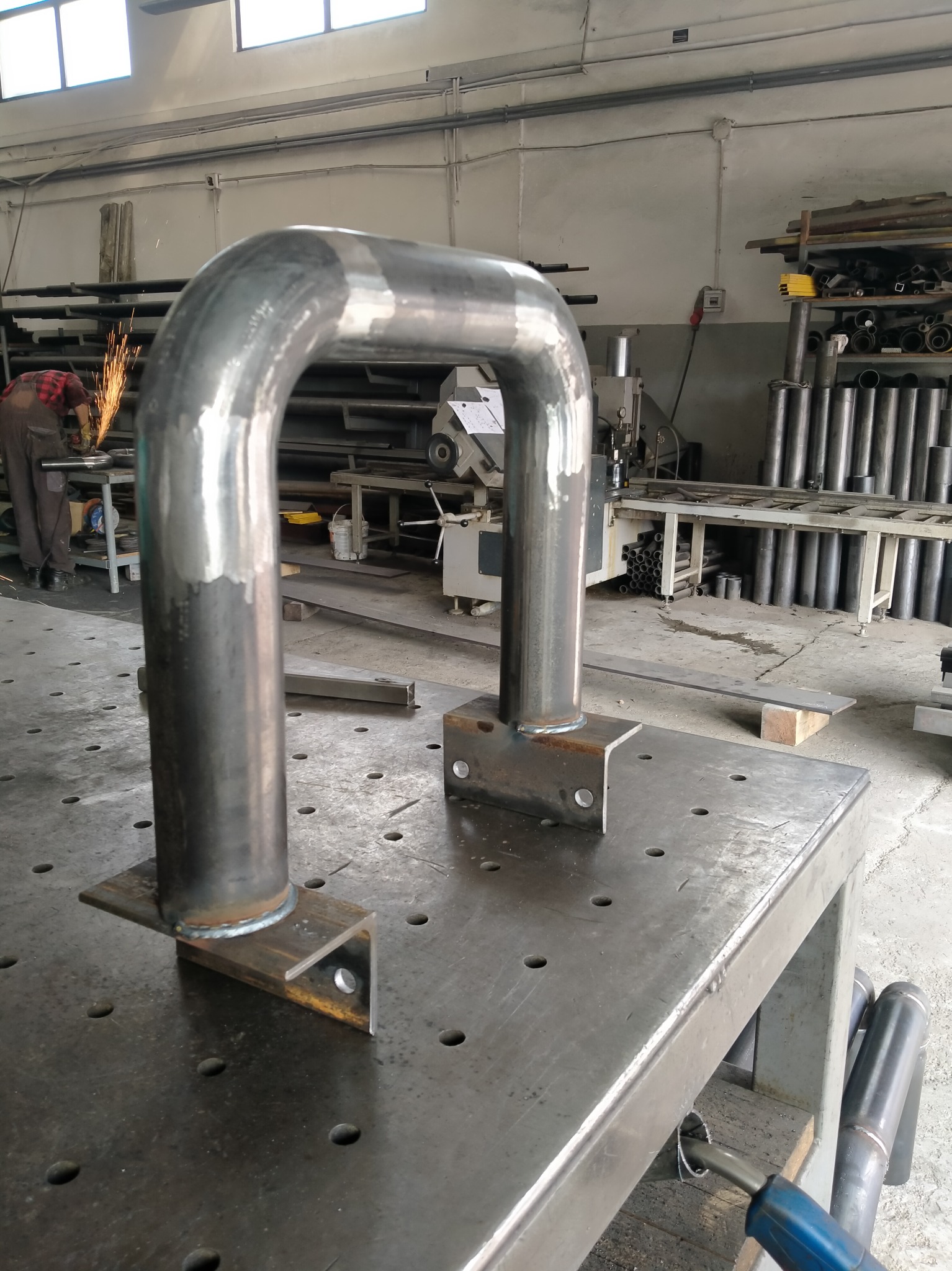 Metalowa konstrukcja w kształcie litery U, spawana do stalowych podstaw, leży na perforowanym stole warsztatowym; w tle iskry spawania i maszyny warsztatowe.