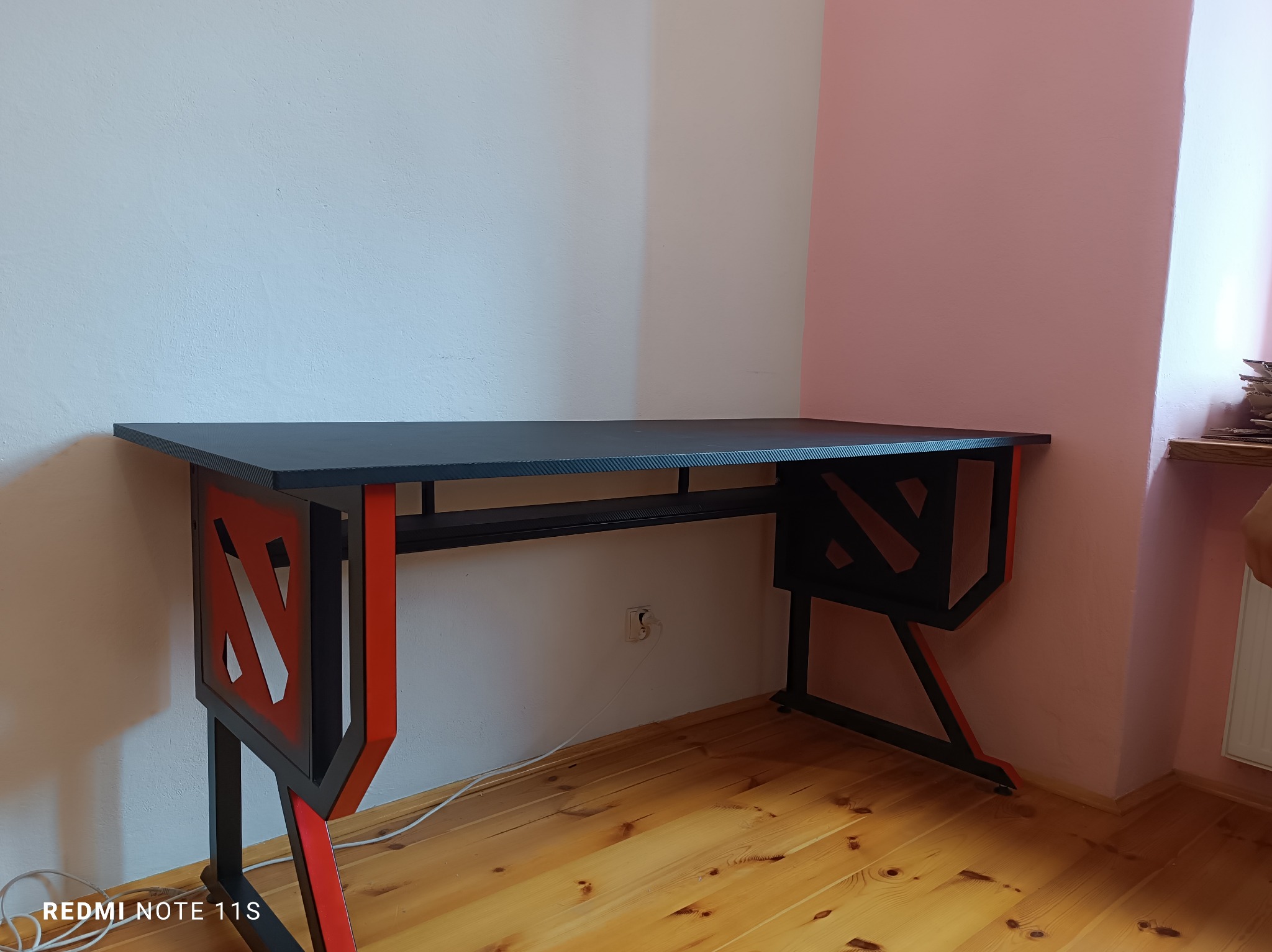 Czarno-czerwone biurko gamingowe z logo Dota 2 na nogach, ustawione w rogu pokoju z drewnianą podłogą i ścianami w odcieniach bieli i pudrowego różu.