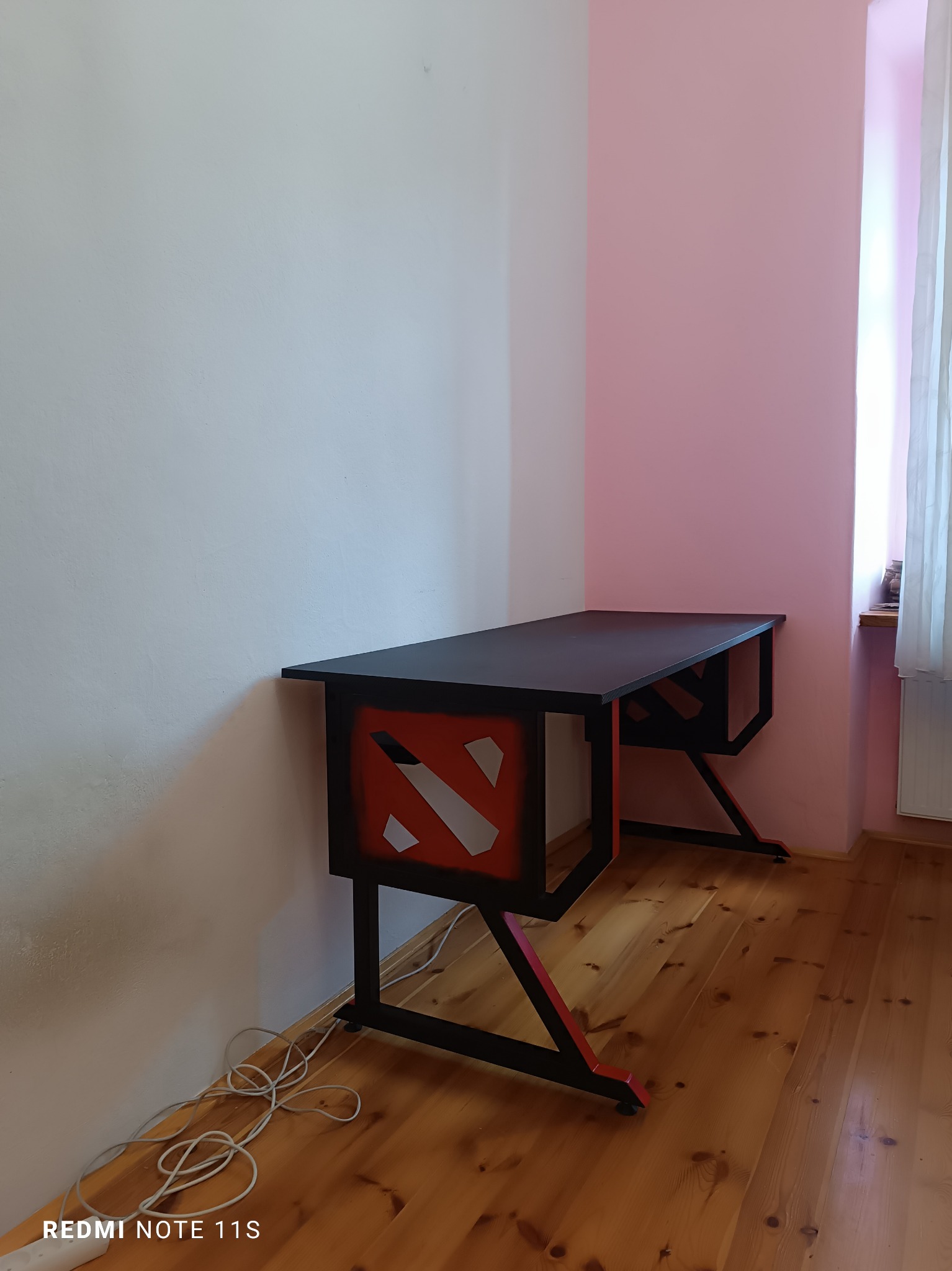 Czarno-czerwone biurko gamingowe z motywem graficznym na tle ściany w dwóch odcieniach różu i drewnianej podłogi.