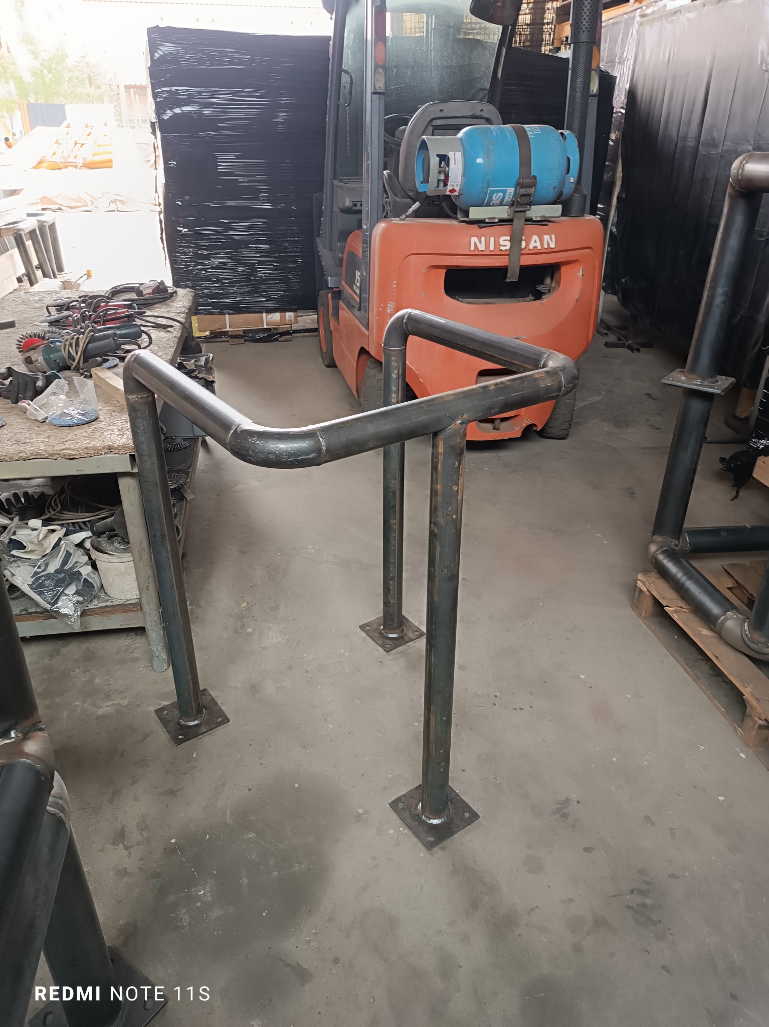 Surowa, spawana konstrukcja metalowa z rur, stojąca na kwadratowych podstawach, w warsztacie, z wózkiem widłowym w tle. Widoczne ślady spawania i niedoskonałości powierzchni.