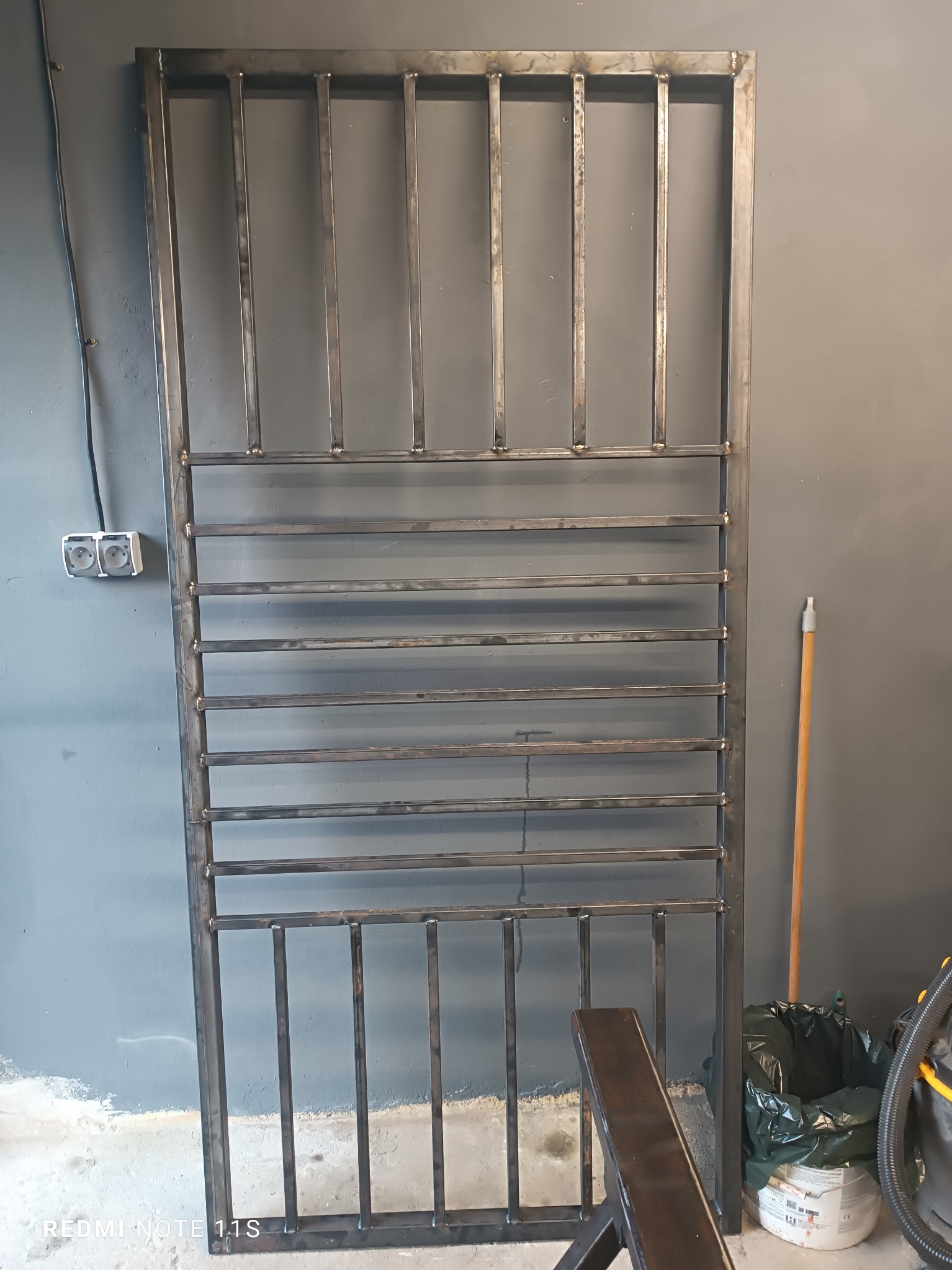 Surowa, metalowa konstrukcja bramy z prostokątnymi prętami, widoczne spawy, oparta o szarą ścianę w warsztacie, obok gniazdko elektryczne i mop.