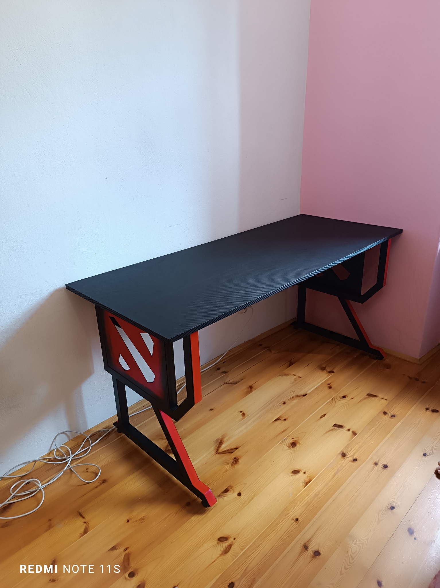 Czarno-czerwone biurko gamingowe z charakterystycznym wzorem na boku, stojące na drewnianej podłodze przy białej i różowej ścianie.