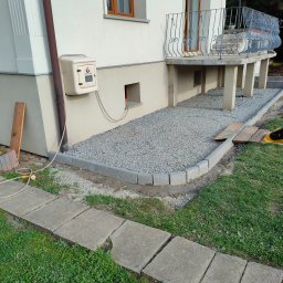 Jonasz Kowala - Obszar pod balkonem domu jednorodzinnego przygotowany z obrzeżami z kostki i wysypany szarą frakcją kamienną, częściowo utwardzony zagęszczarką.