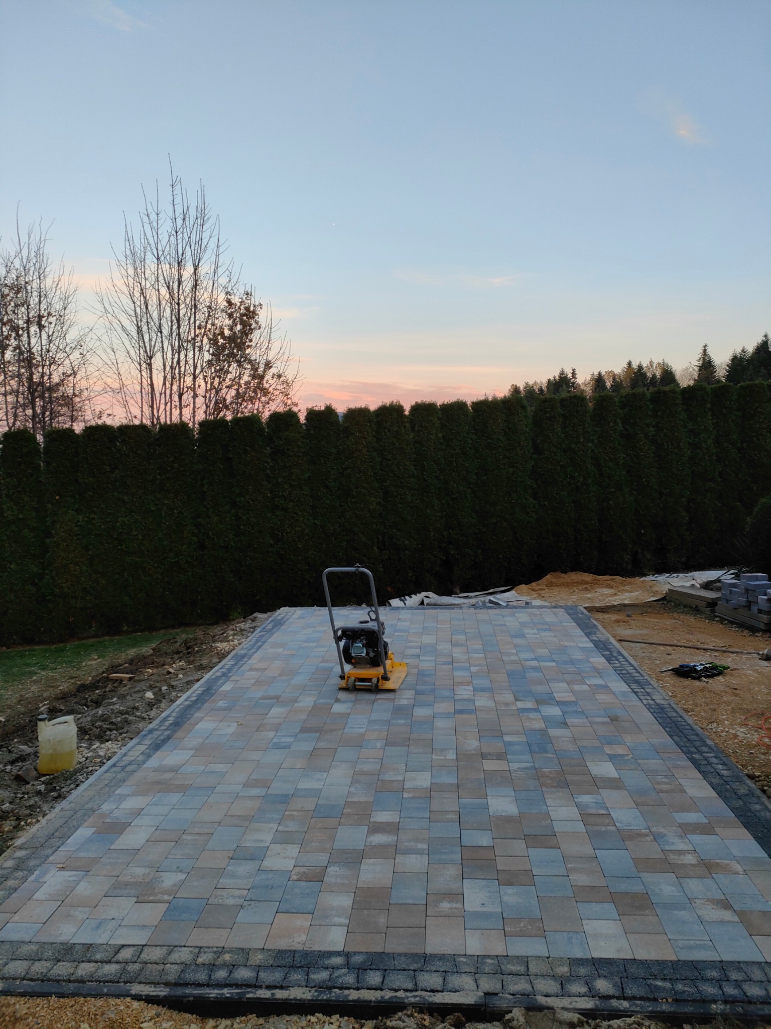 Wibrująca zagęszczarka stoi na częściowo ułożonej kostce brukowej w odcieniach szarości, beżu i błękitu, otoczonej zielonym żywopłotem i drzewami o zachodzie słońca.