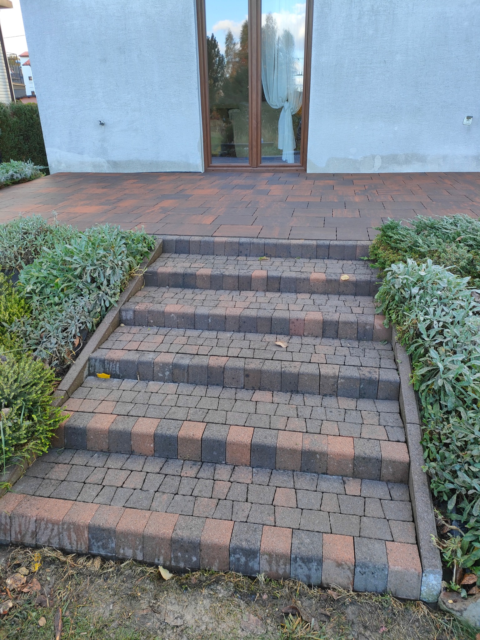 Schody z kostki brukowej dwukolorowej, prowadzące do domu z jasną elewacją i brązowymi oknami, z roślinami po bokach.