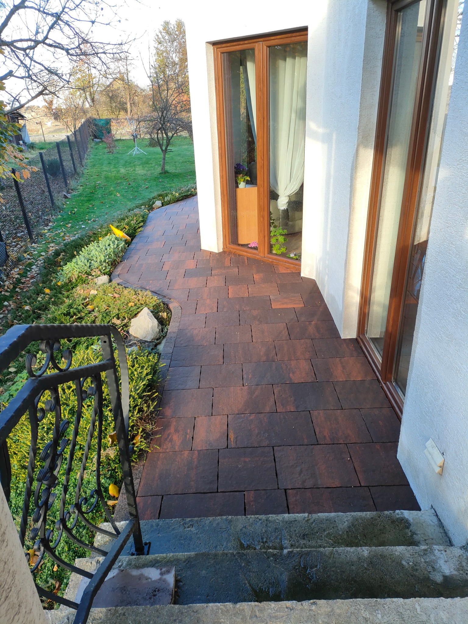 Brązowa kostka brukowa ułożona na ścieżce przy domu, widok z góry ze schodów z ozdobną, metalową balustradą, w tle trawnik z drzewami i ogrodzeniem.