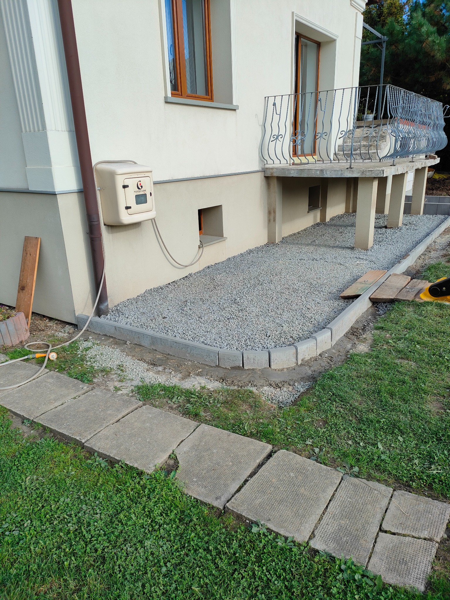 Obszar pod balkonem domu jednorodzinnego przygotowany z obrzeżami z kostki i wysypany szarą frakcją kamienną, częściowo utwardzony zagęszczarką.