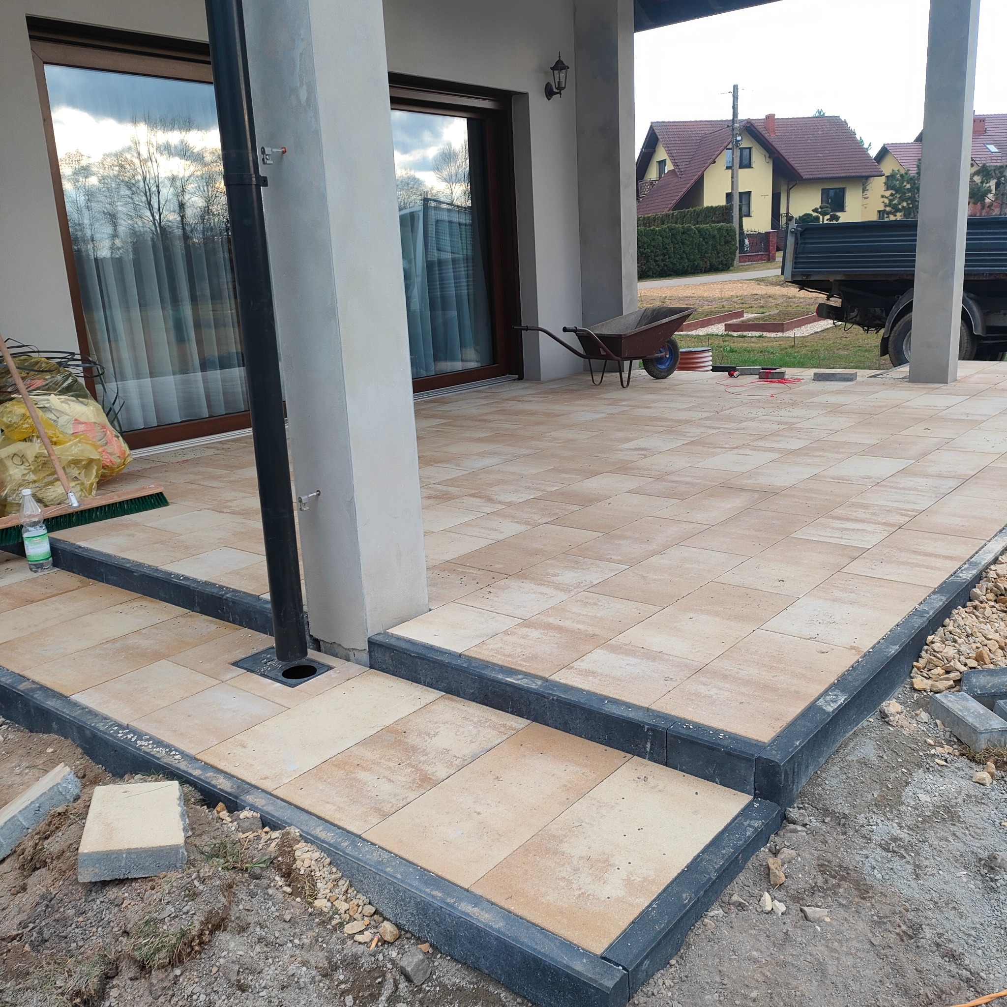Nowo ułożona, beżowa kostka brukowa na tarasie domu jednorodzinnego z czarnymi obrzeżami; widoczny fragment elewacji, rynna spustowa, taczka w tle i przygotowane kamienie do wykończenia.
