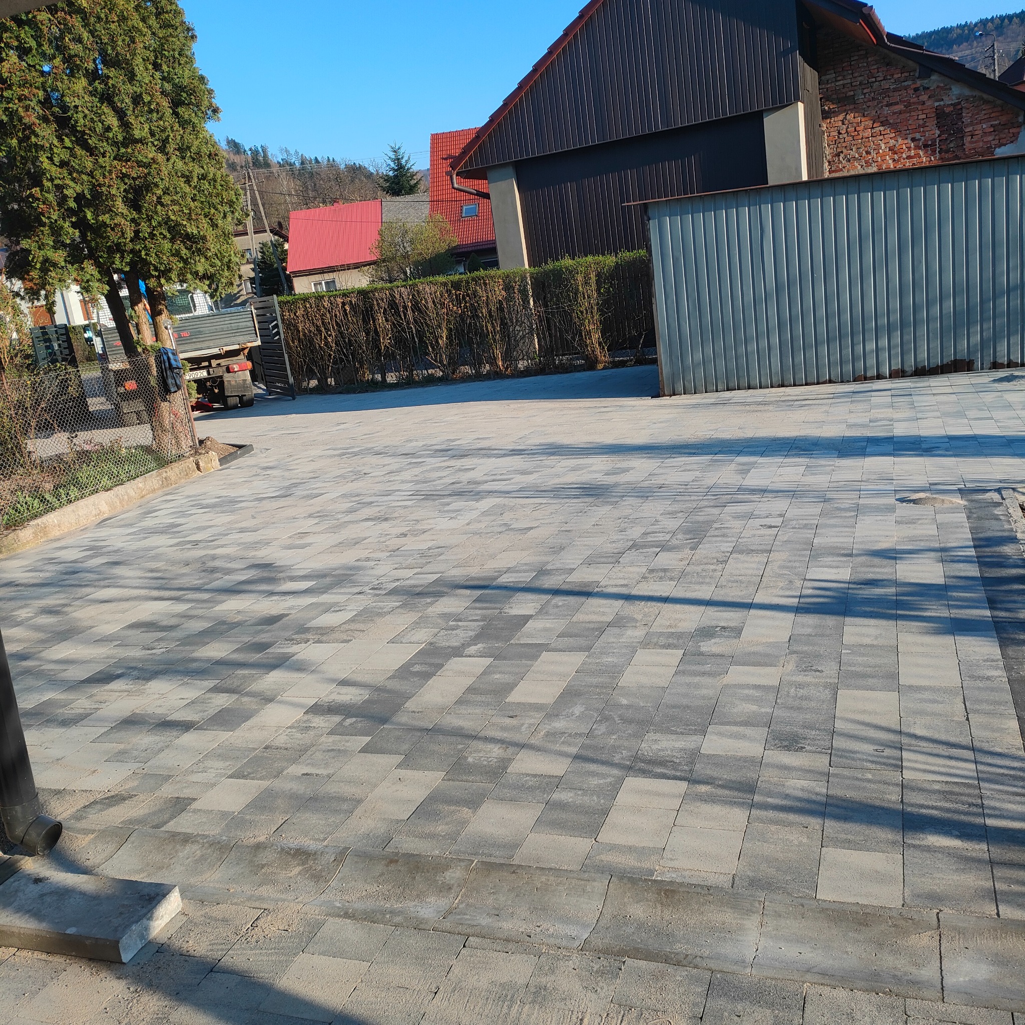 Ułożona szara kostka brukowa na posesji z widocznym drzewem i budynkami w tle, oświetlona popołudniowym słońcem rzucającym cienie.