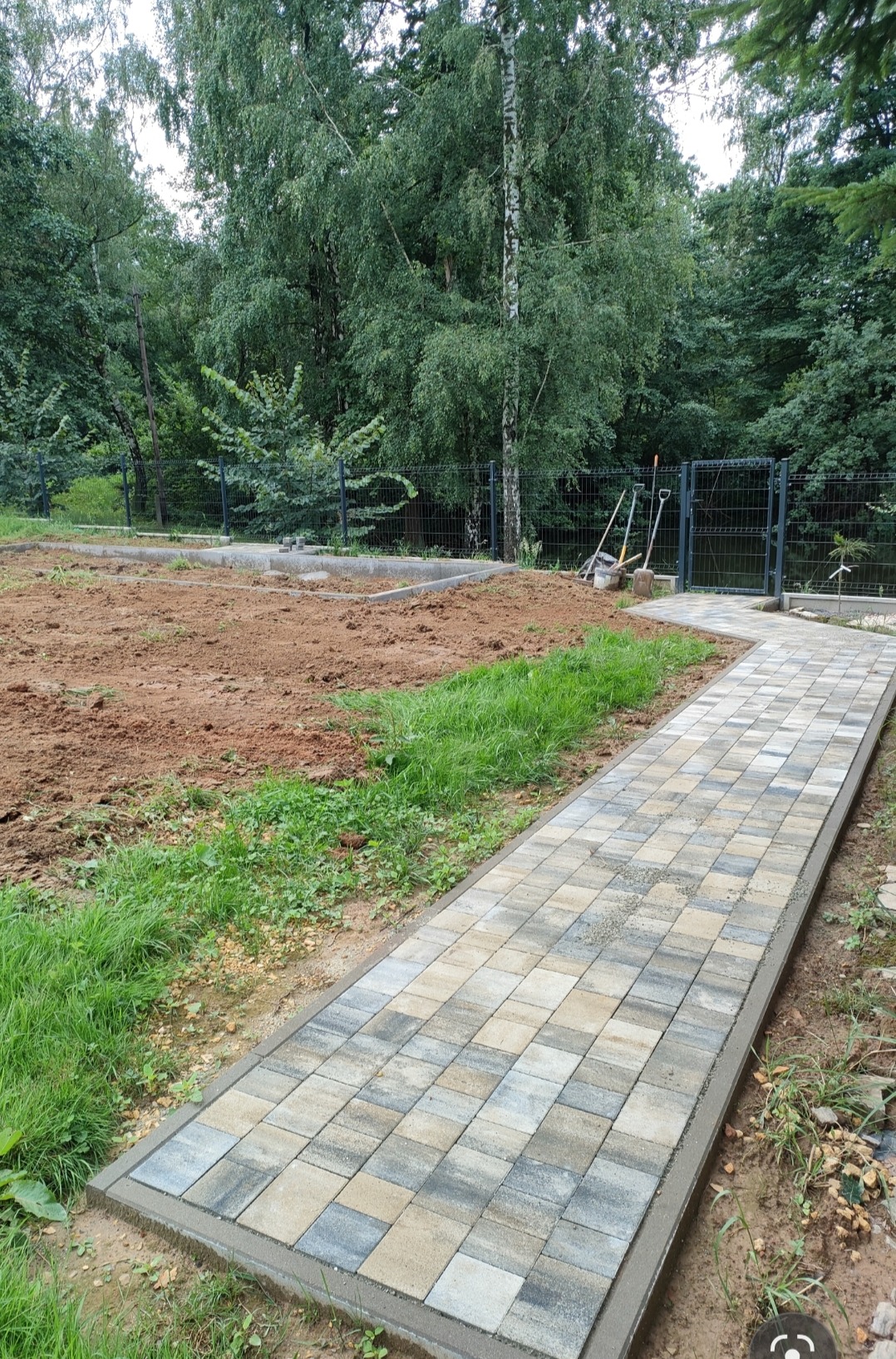 Ścieżka z kostki brukowej prowadząca do furtki w ogrodzeniu, obok przygotowany teren pod przyszły ogród, widoczne narzędzia ogrodnicze oparty o ogrodzenie.
