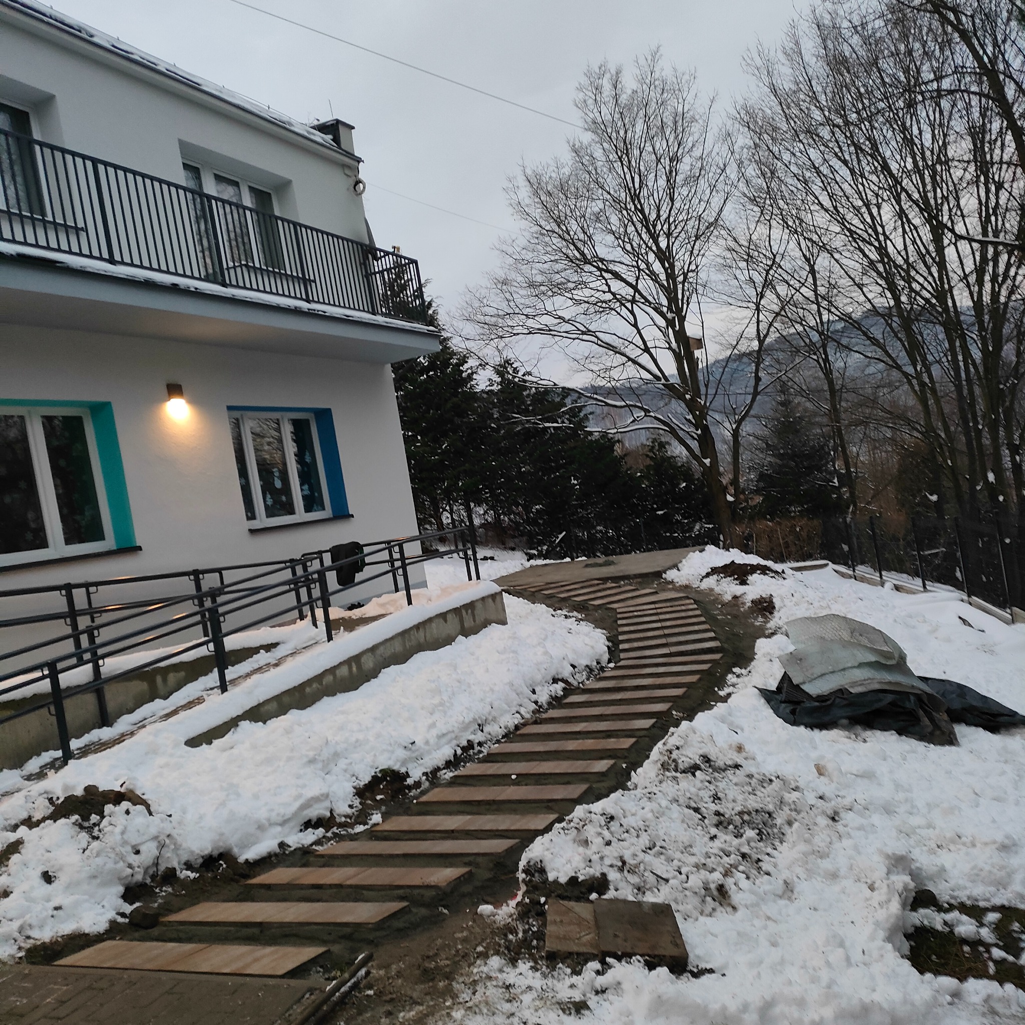 Schody z płyt kamiennych prowadzące do budynku z barierkami, otoczone śniegiem i drzewami na pochmurnym tle.