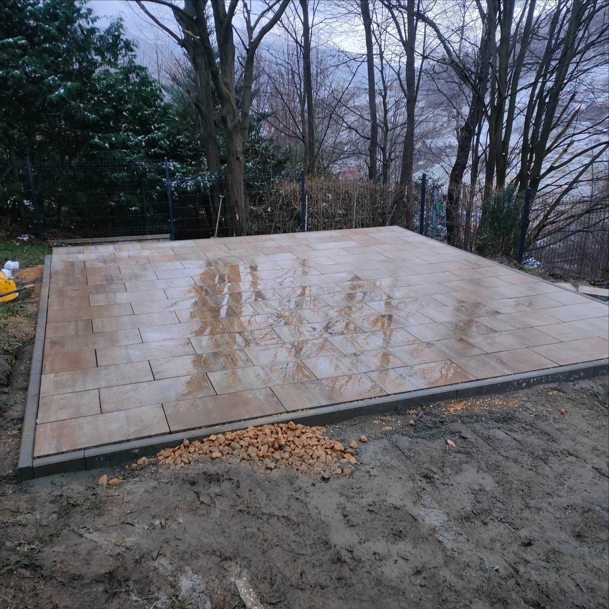 Wykończona powierzchnia z beżowej kostki brukowej, obrzeża z szarego betonu, widoczne brązowe kamyczki na ziemi, w tle drzewa bez liści i fragmenty zieleni, lekko pochmurna pogoda.