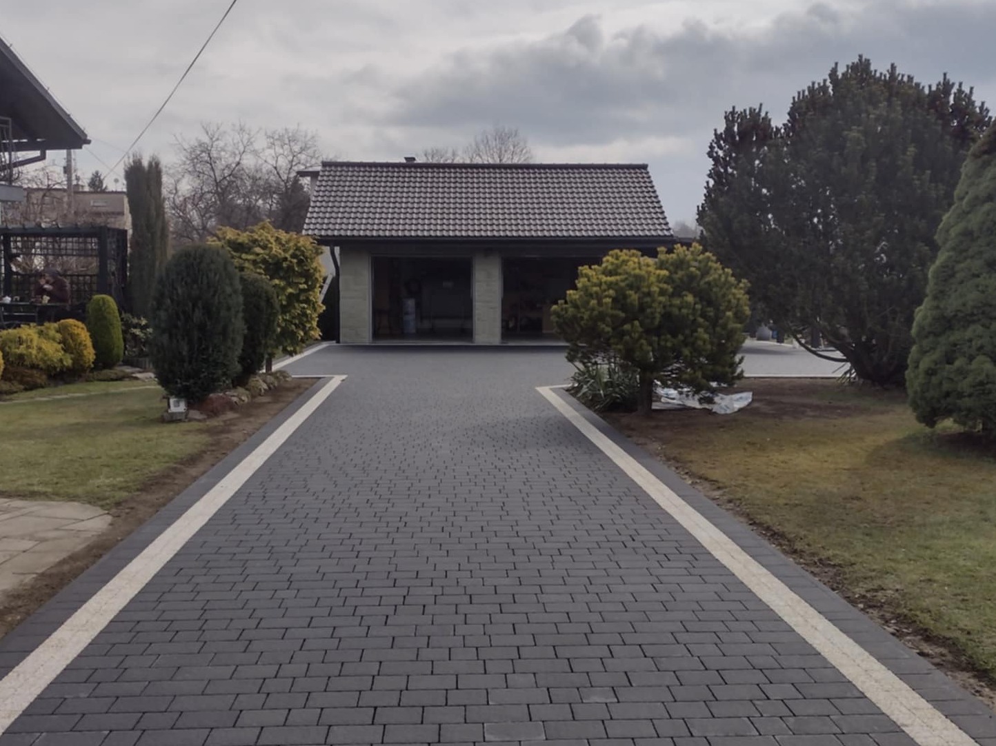 Nowo ułożony, ciemnoszary podjazd z kostki brukowej z jasnymi obrzeżami prowadzący do garażu w otoczeniu zieleni w pochmurny dzień.